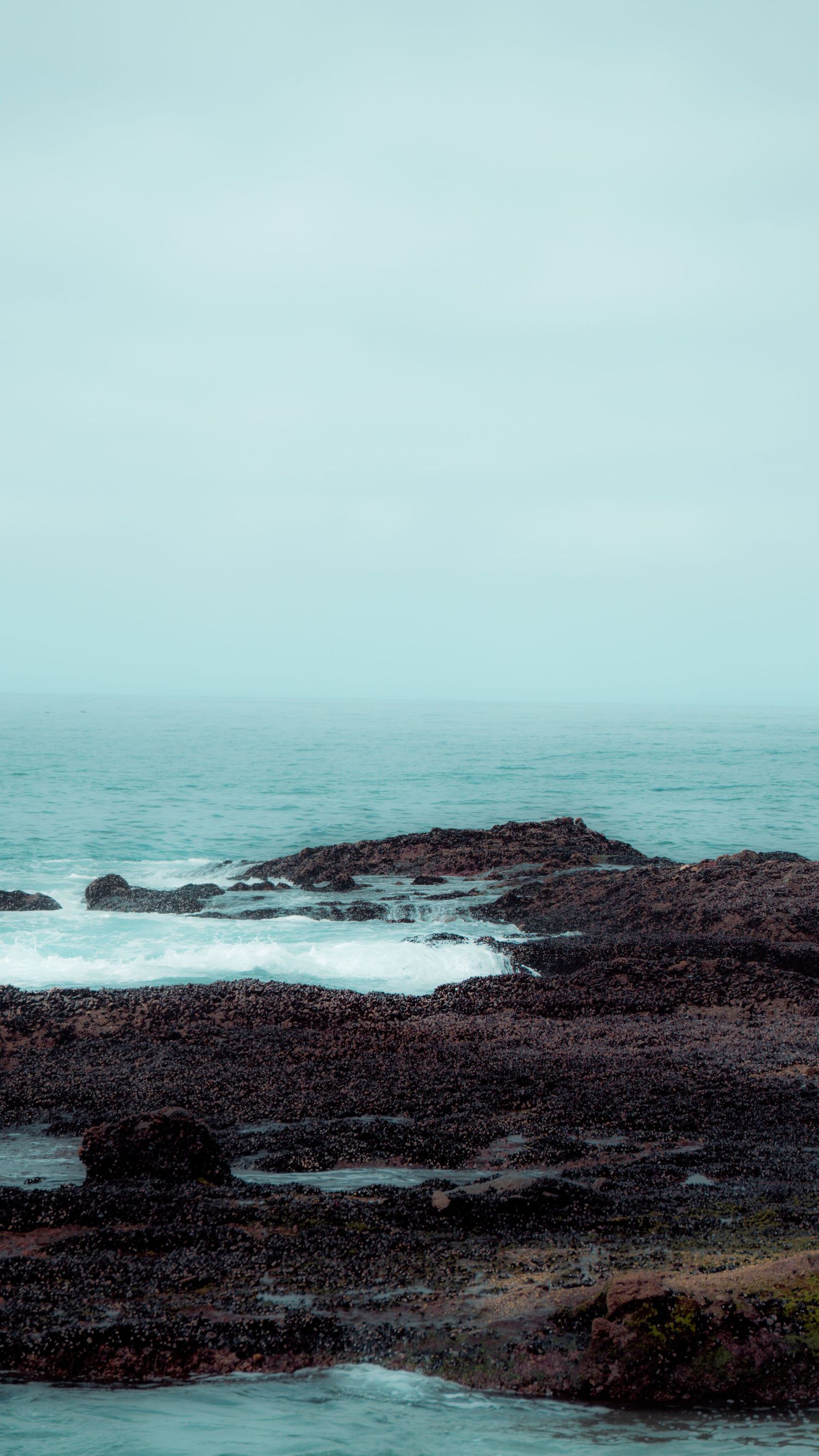 Rugged volcanic coastline with waves breaking against dark rocks under a hazy turquoise sky.