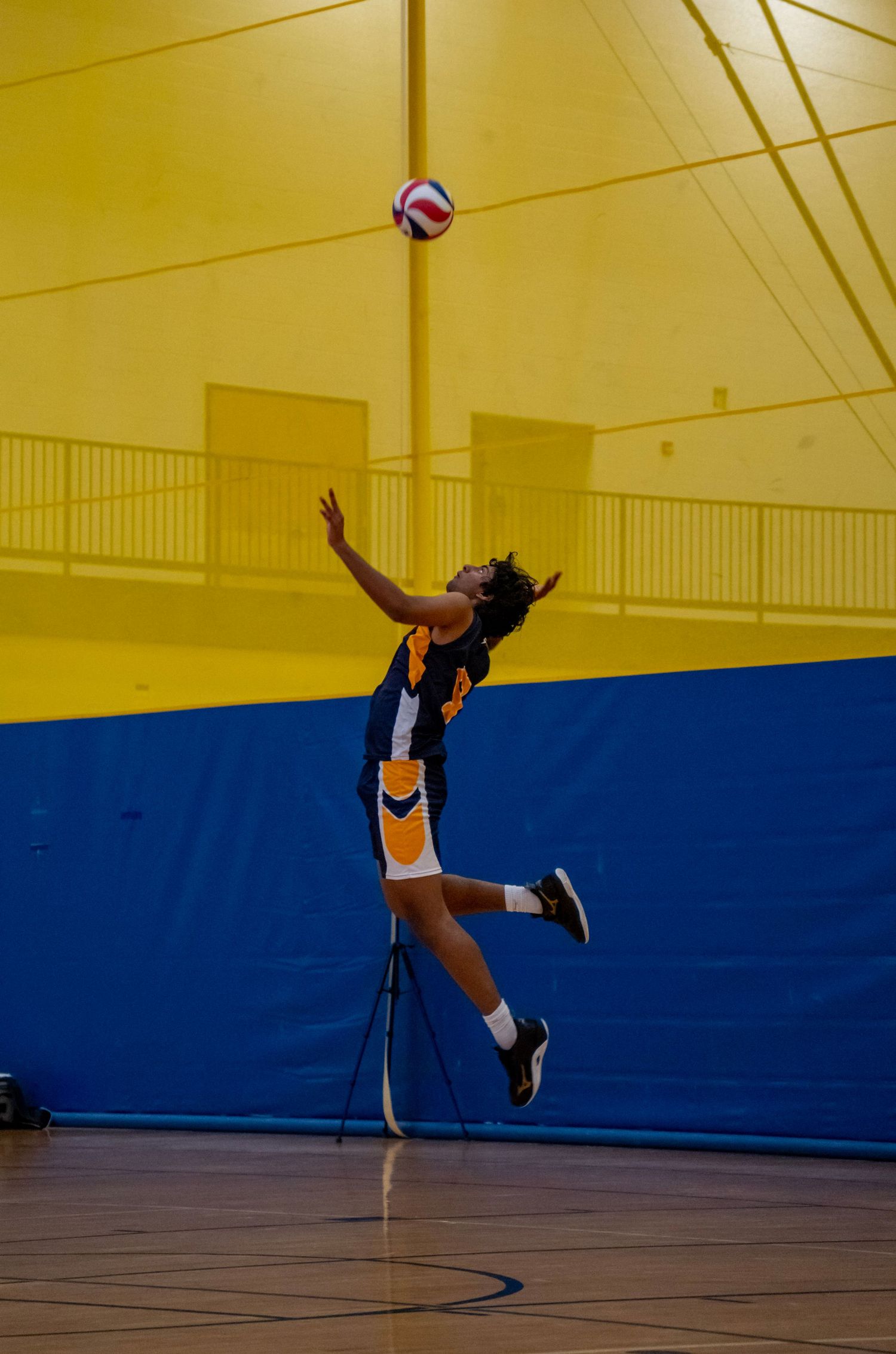 A volleyball player leaps high to serve in a yellow and blue gymnasium.