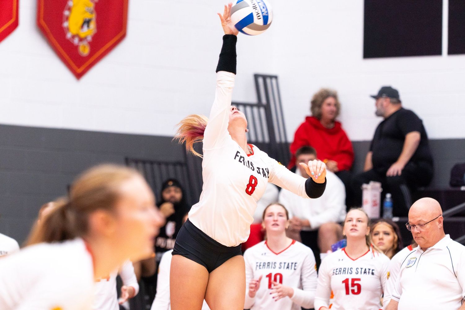 Volleyball player in a white Ferris State jersey number 8 leaps to spin a serve as teammates and spectators watch from the sideline.