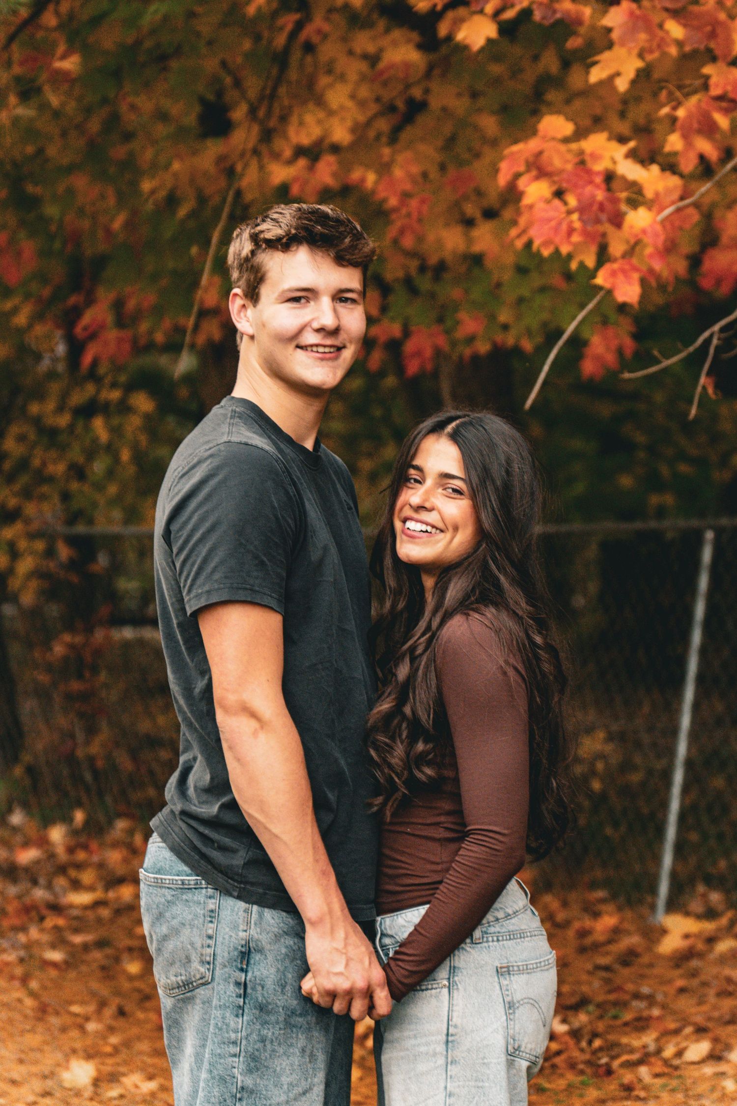 Smiling young couple holding hands and posing together outdoors beneath vibrant orange and red autumn foliage in a fall park setting.