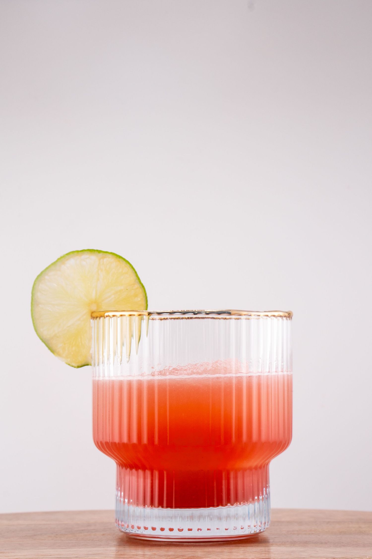 Red cocktail in ribbed glass tumbler garnished with lime wheel.