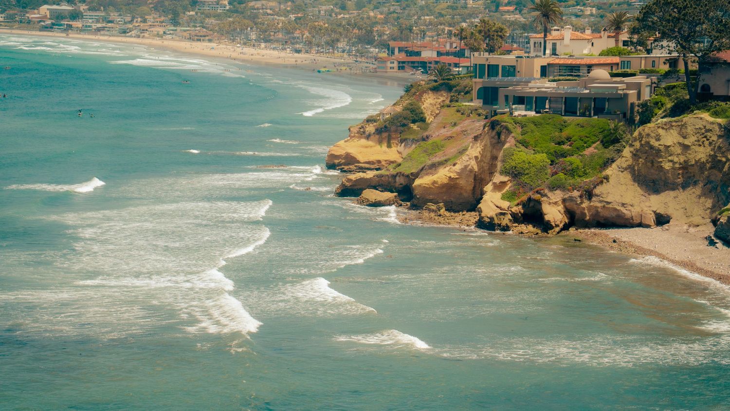 Turquoise waves roll into a scenic coastal cove surrounded by residential cliffs.