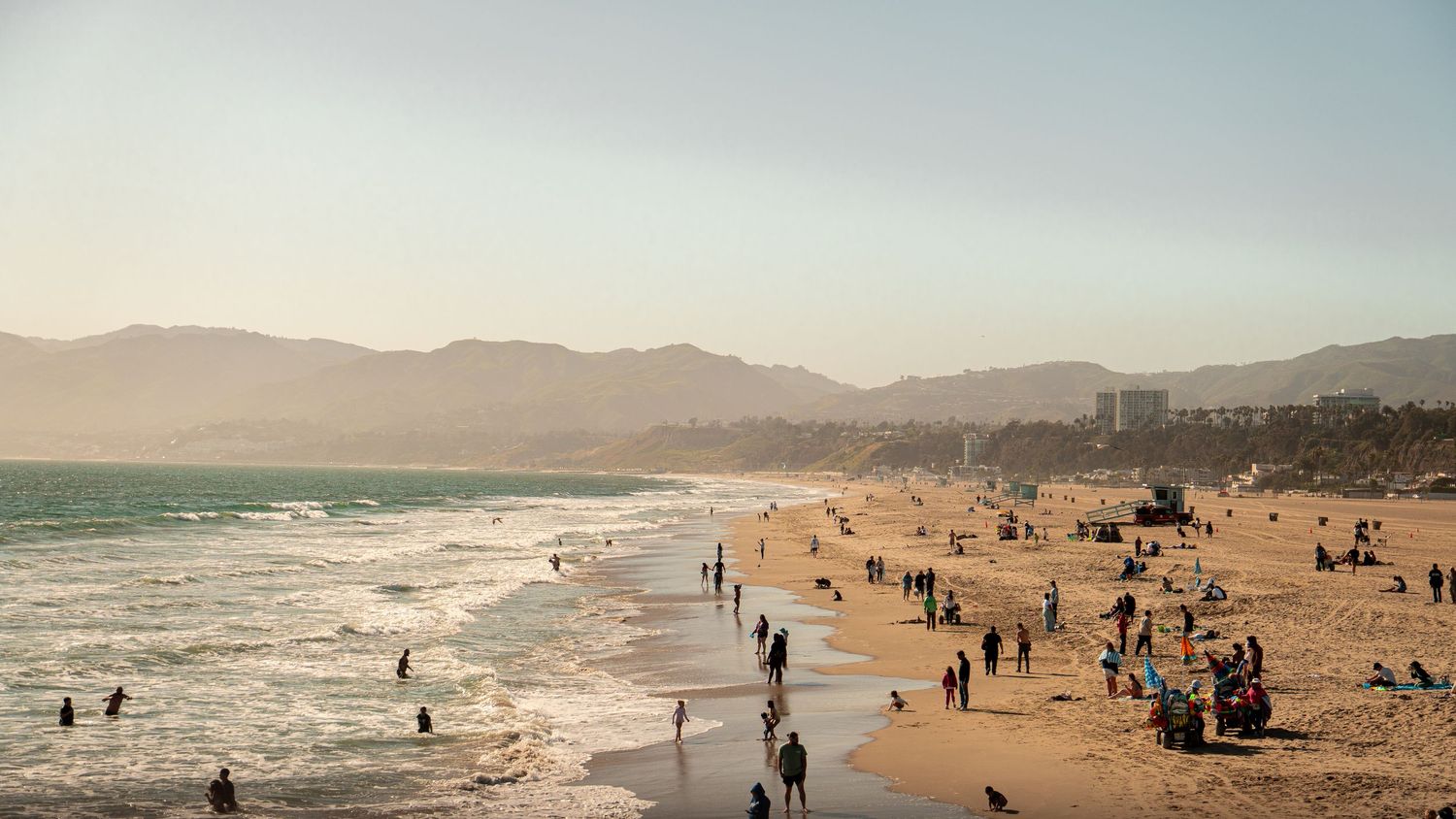 Wide panoramic view of a busy beach with mountains in the background during golden hour.