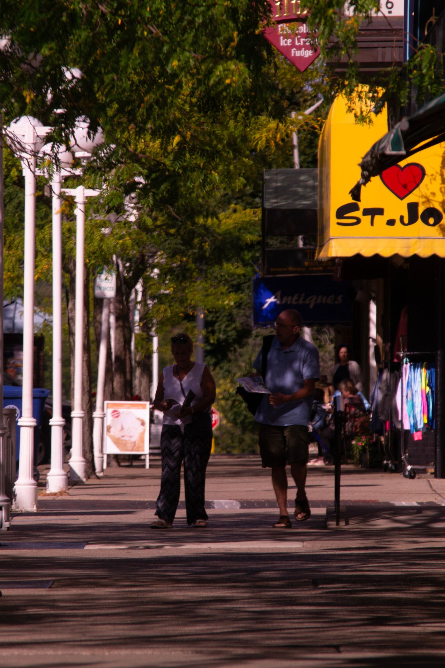 People walking on a shaded sidewalk with street lamps and storefronts in an urban setting.