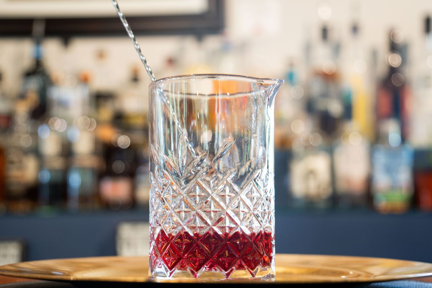 Cut crystal glass containing red syrup with mixing spoon against blurred bar background.