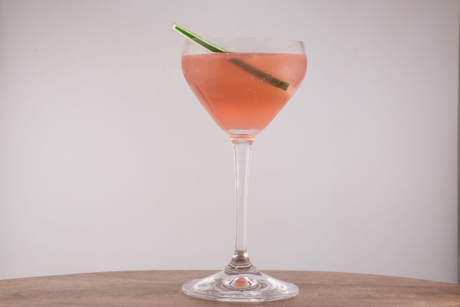 Blush-colored cocktail in coupe glass garnished with lemongrass on wooden table.