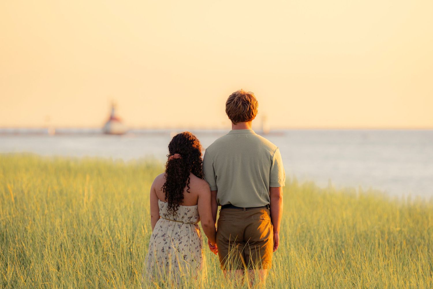 Couple holding hands while walking through a sunlit meadow at golden hour along the coastline.