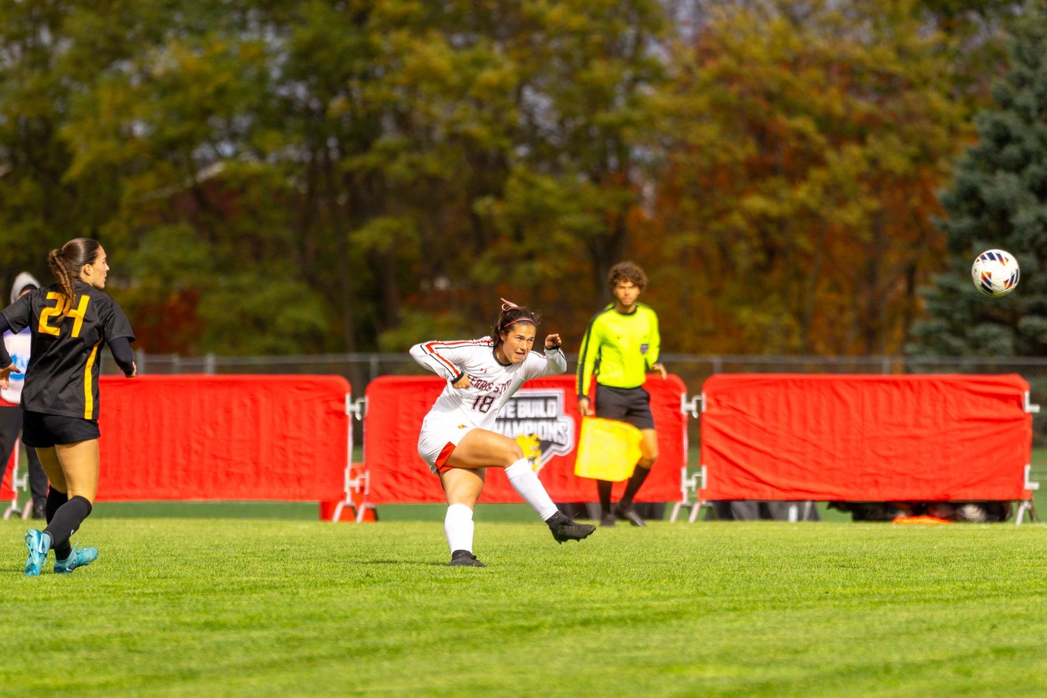 Soccer players compete on a grass field during a match with fall foliage visible in the background.