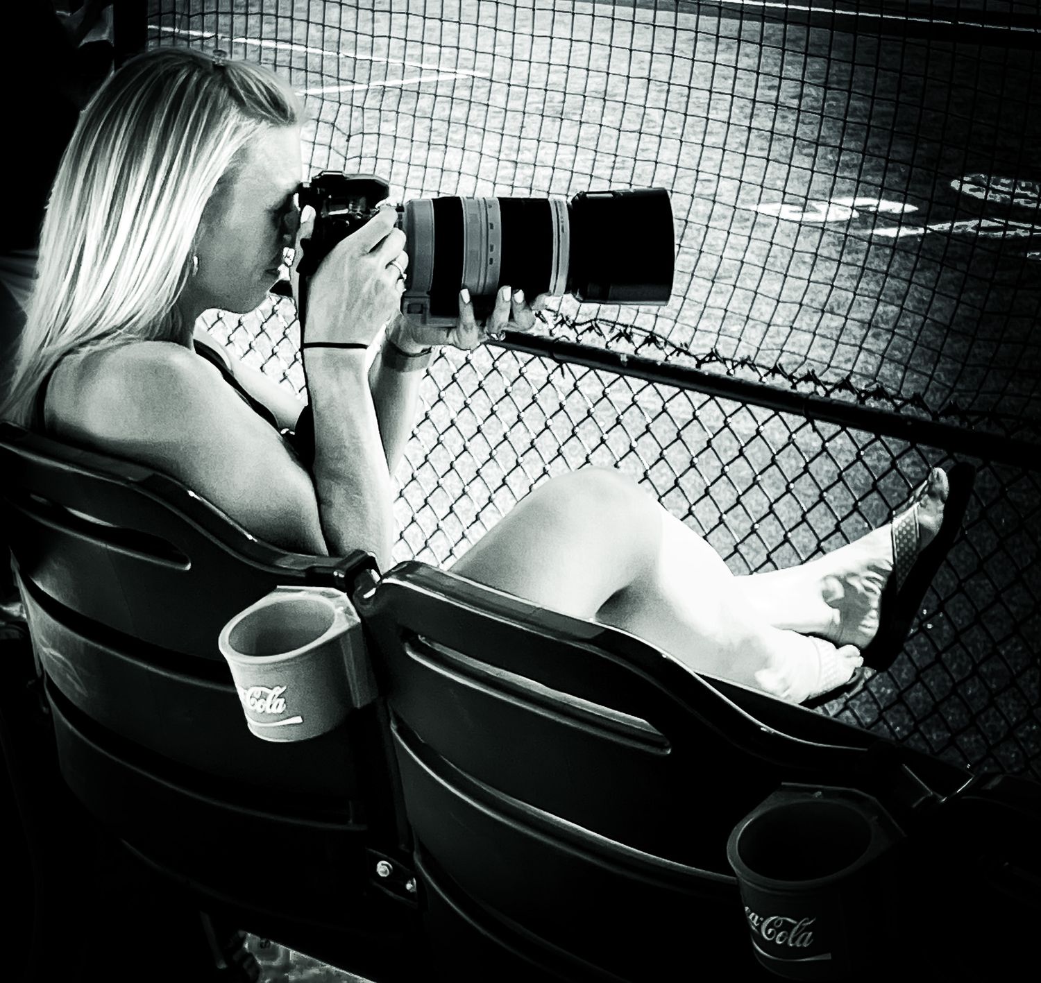 Black and white photo of a photographer relaxing barefoot with camera at a baseball game behind protective netting.