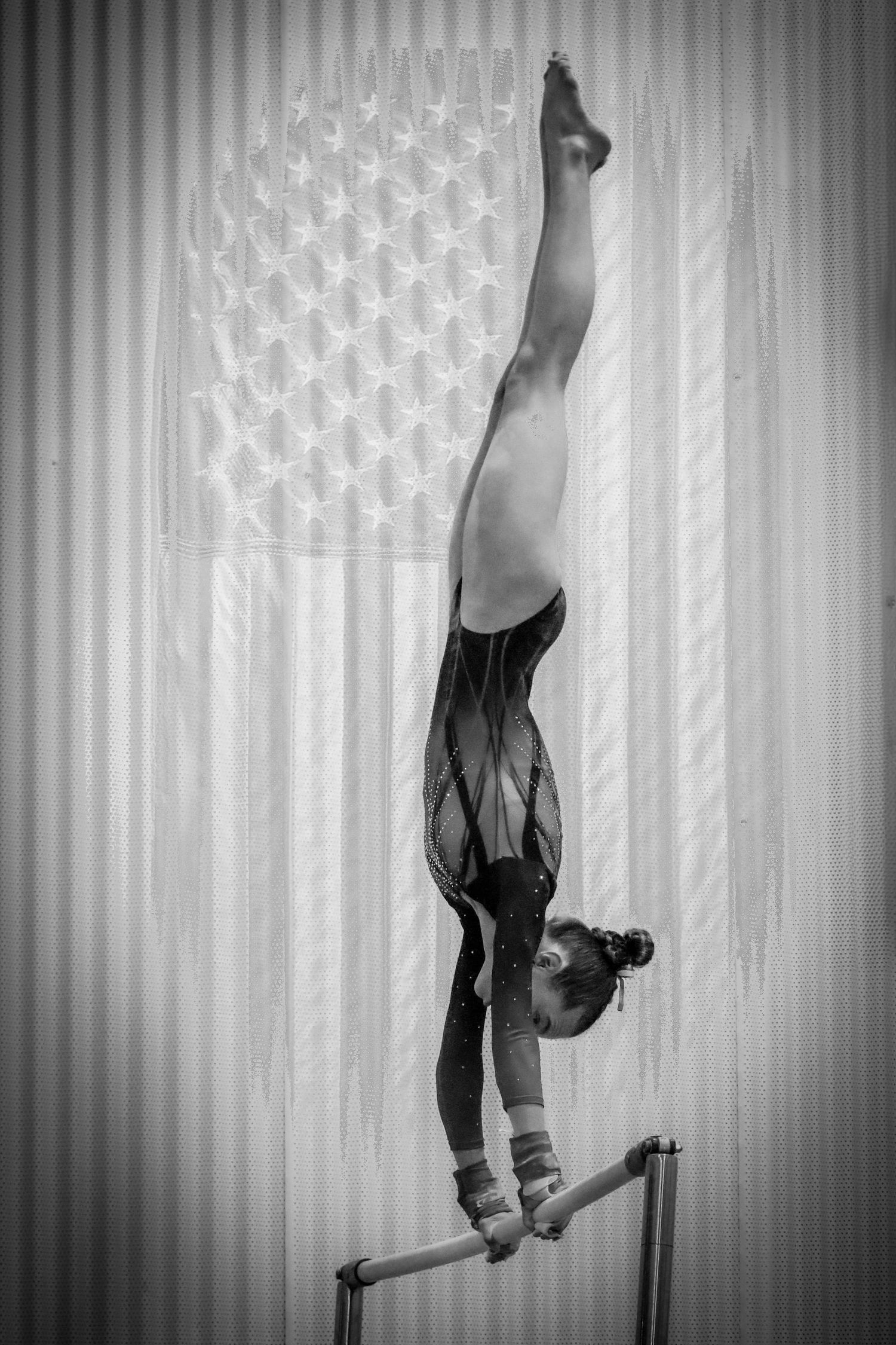 A gymnast performs an giant (inverted handstand) on uneven bars with the American flag backdrop in black and white photography.