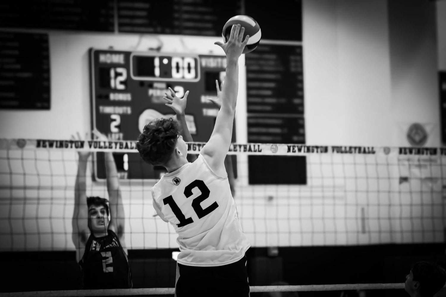 Volleyball player in jersey number 12 leaps high to spike the ball over the net during indoor game.