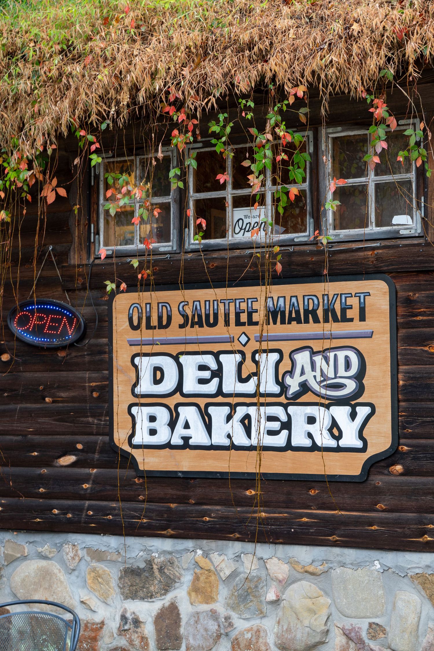 A sign displaying the name of a local cafe with greenery falling into it from the roof and an open sign lit up beside it.
