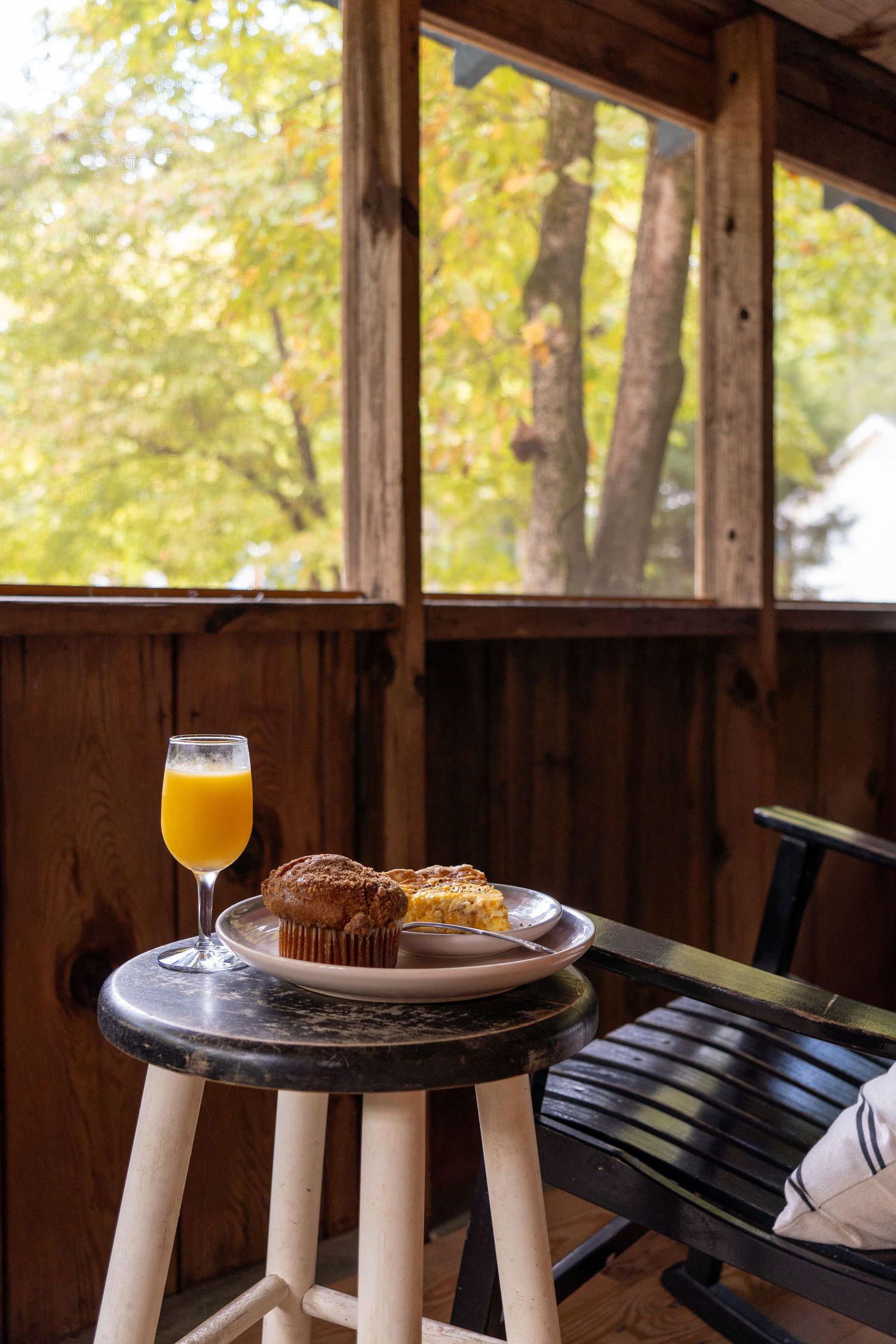 A plate with a muffin and a glass of orange juice sits on a small round table beside a black rocking chair on a wooden porch, with leafy