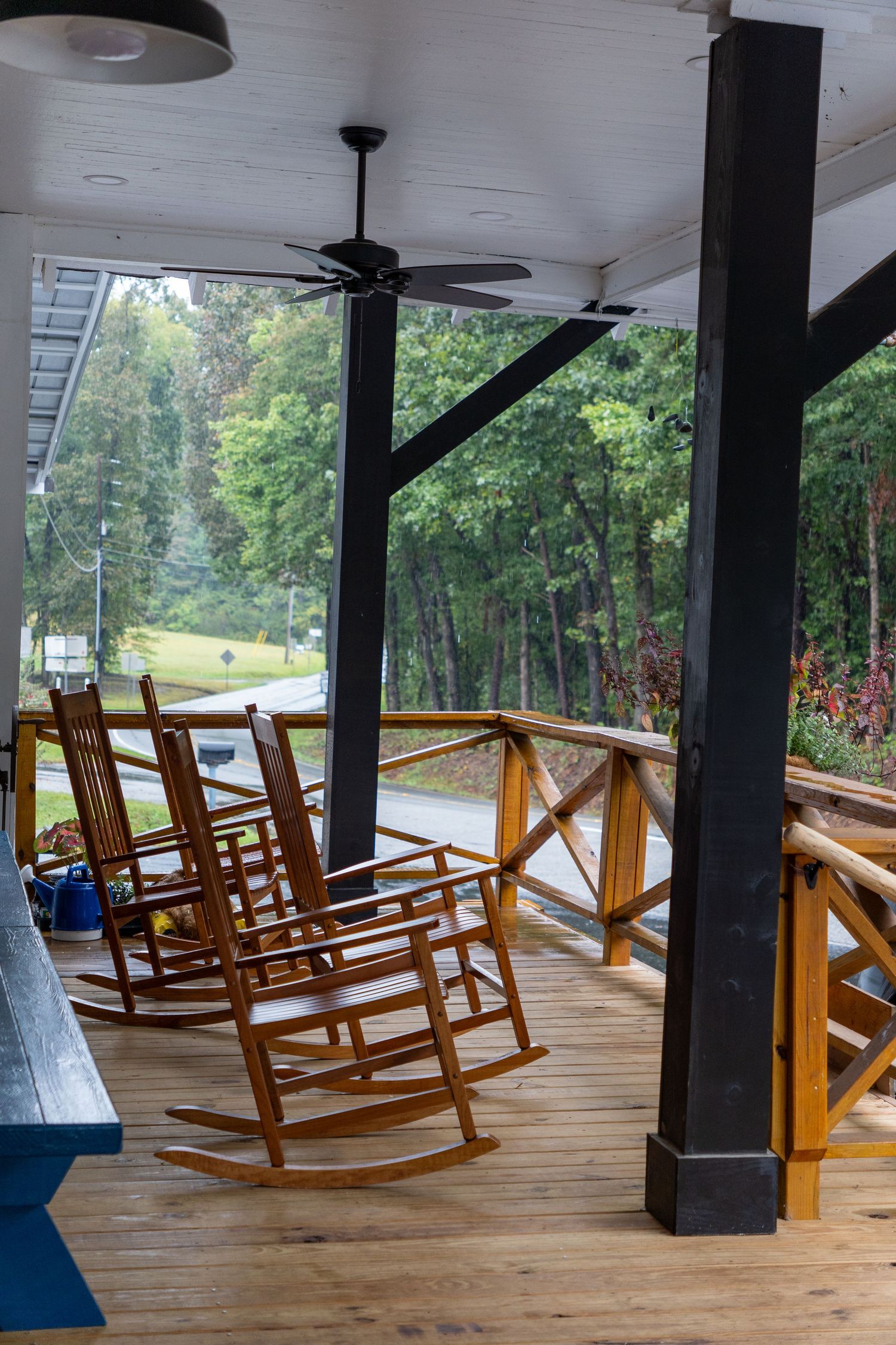 Three wooden rocking chairs sit on a covered porch with wooden railings, overlooking a tree-lined road. The scene is peaceful