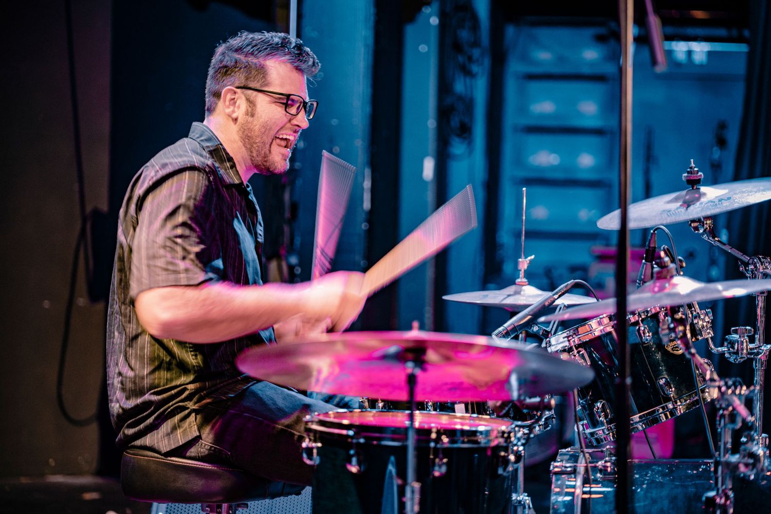 Drummer performs behind full kit with colorful stage lighting and dramatic shadows.