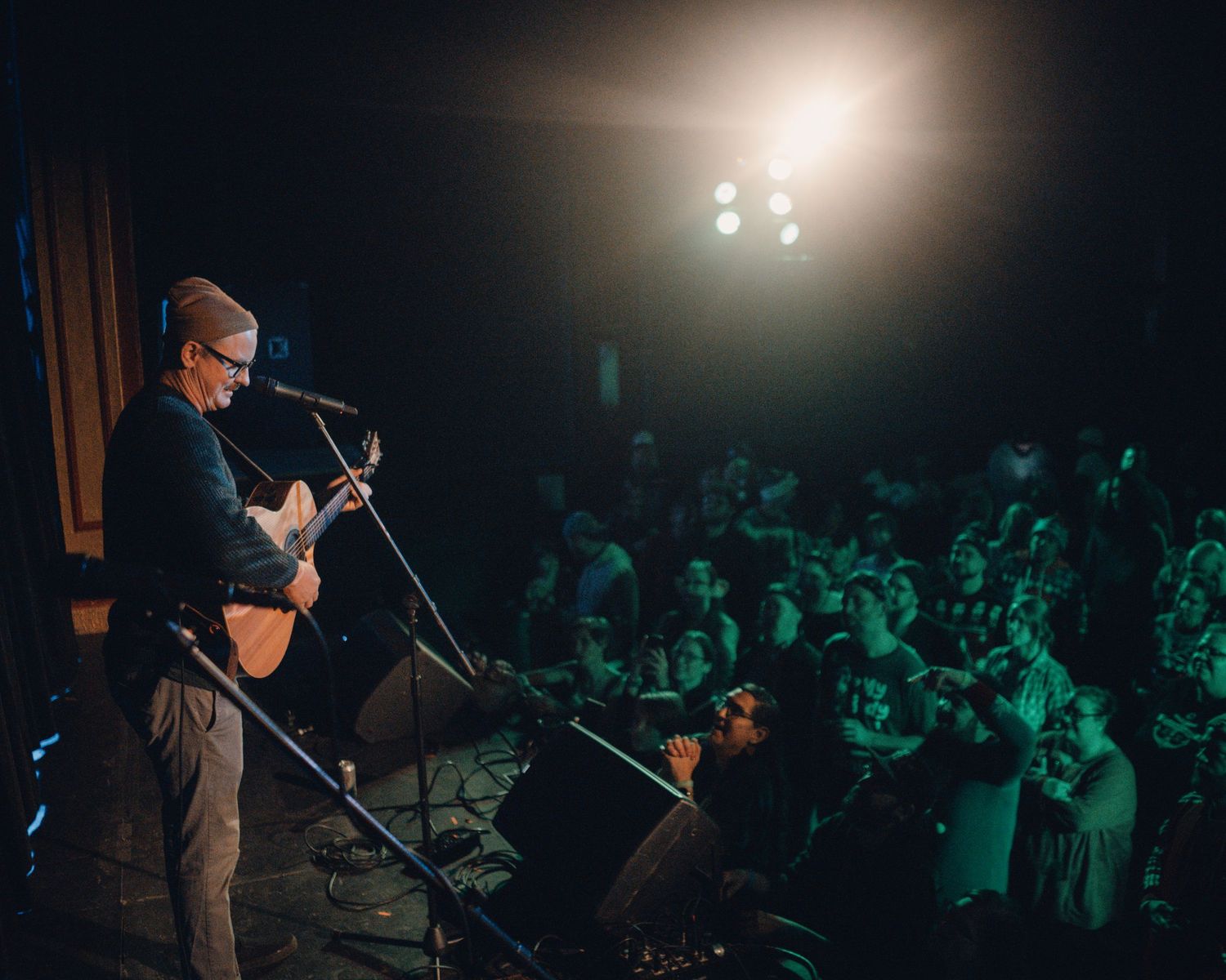 Performer on stage illuminated by spotlight with green-tinted audience in background during live concert.