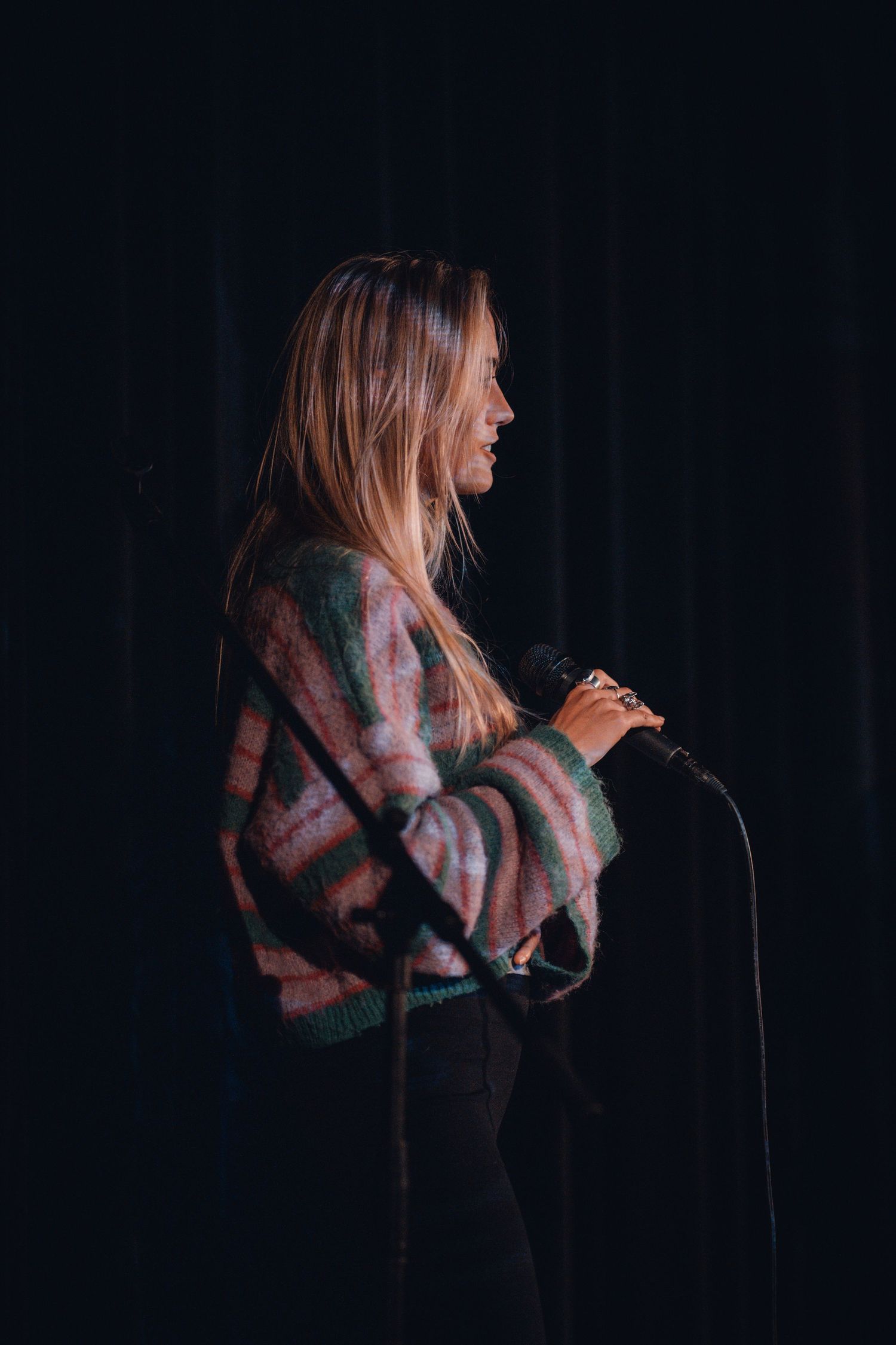 Singer in patterned sweater performing on dimly lit stage with microphone.
