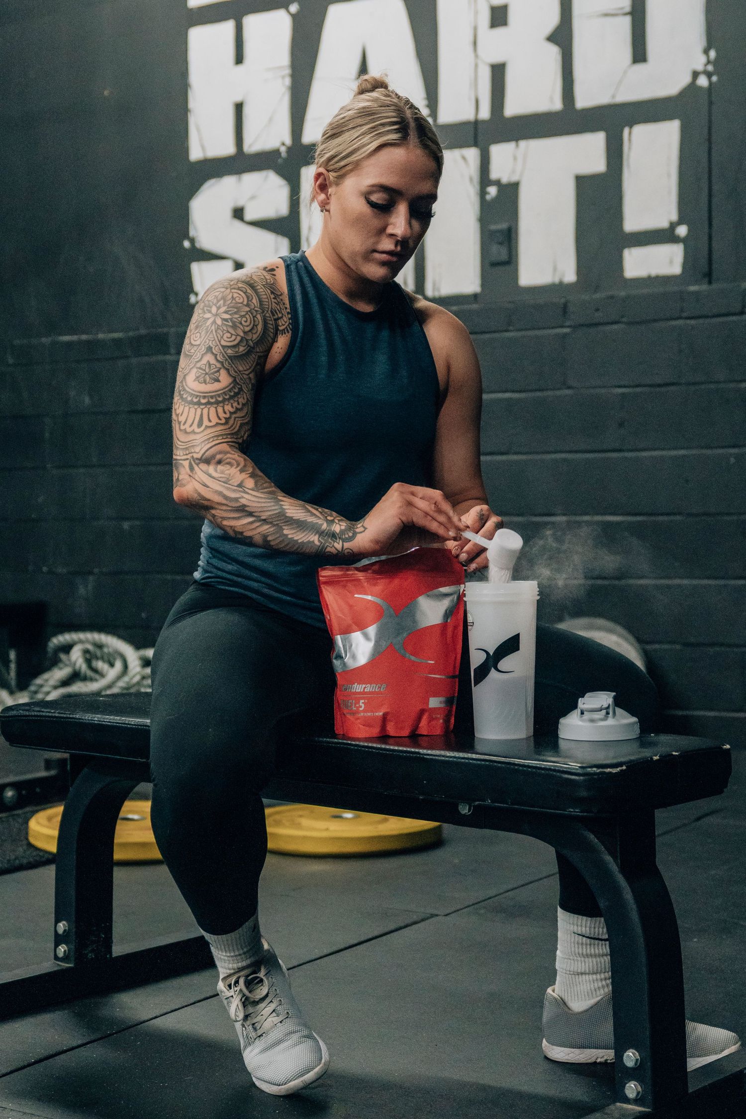 Athlete in black tank top preparing supplements at bench in dark gym setting.