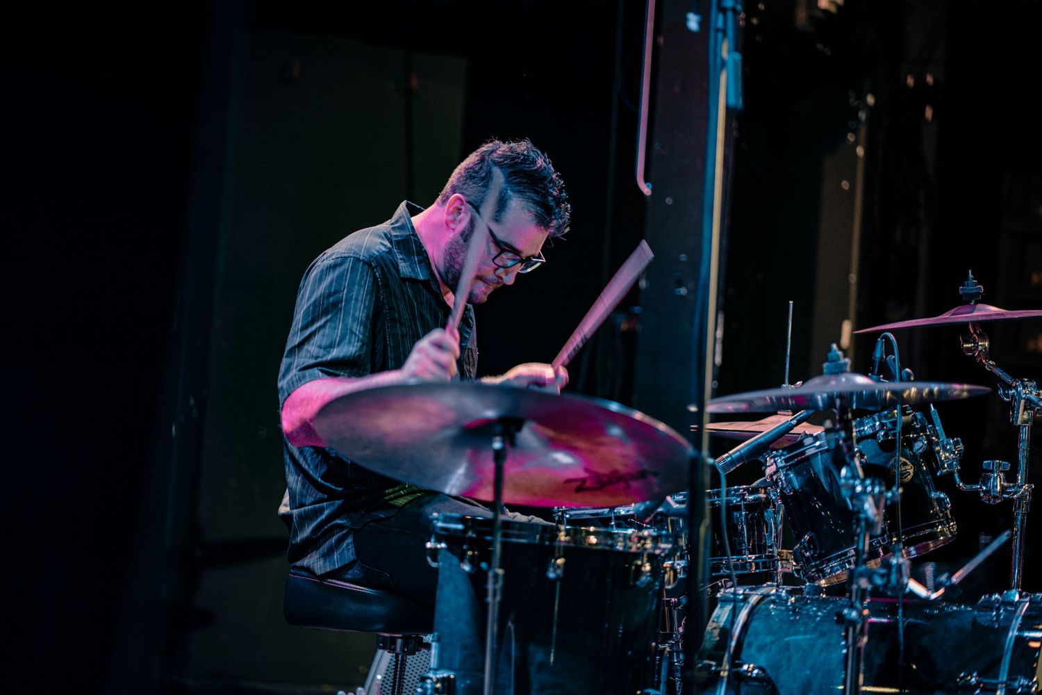 Drummer plays energetically behind kit with dramatic stage lighting creating silhouette effect.