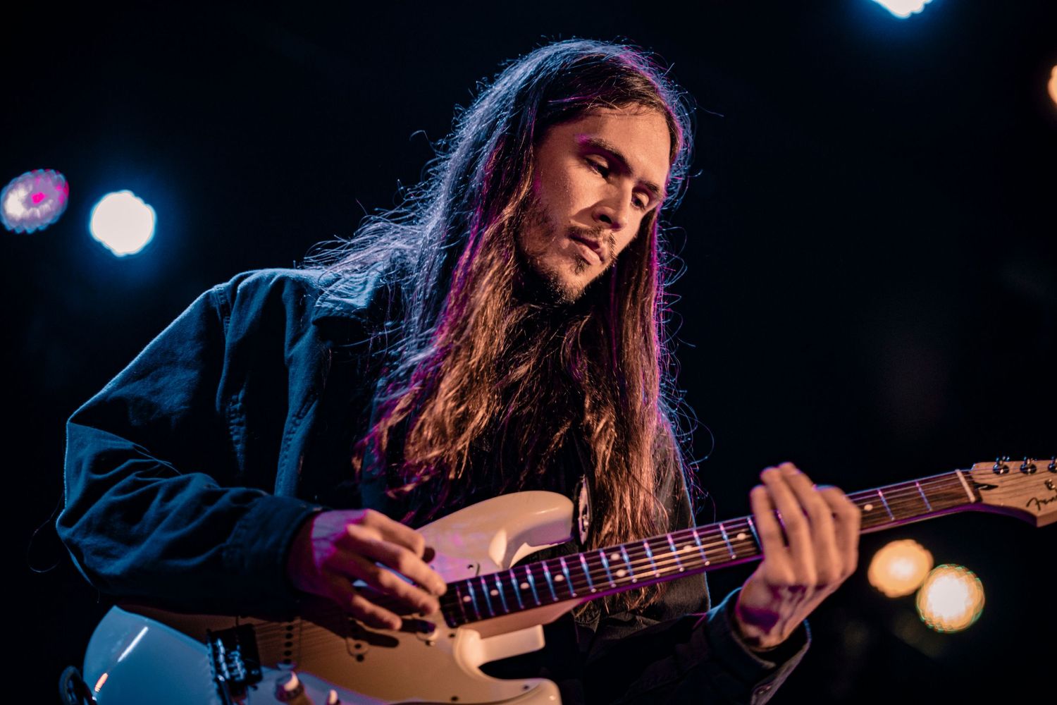 Guitarist plays passionately on dark stage with atmospheric lighting and bokeh effects.