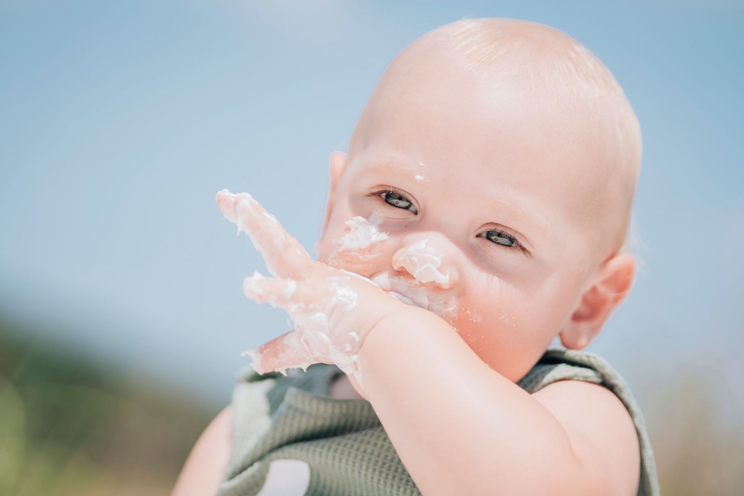 Baby wearing green outfit with white cream or frosting smeared on face smiles during sunny day outdoors.