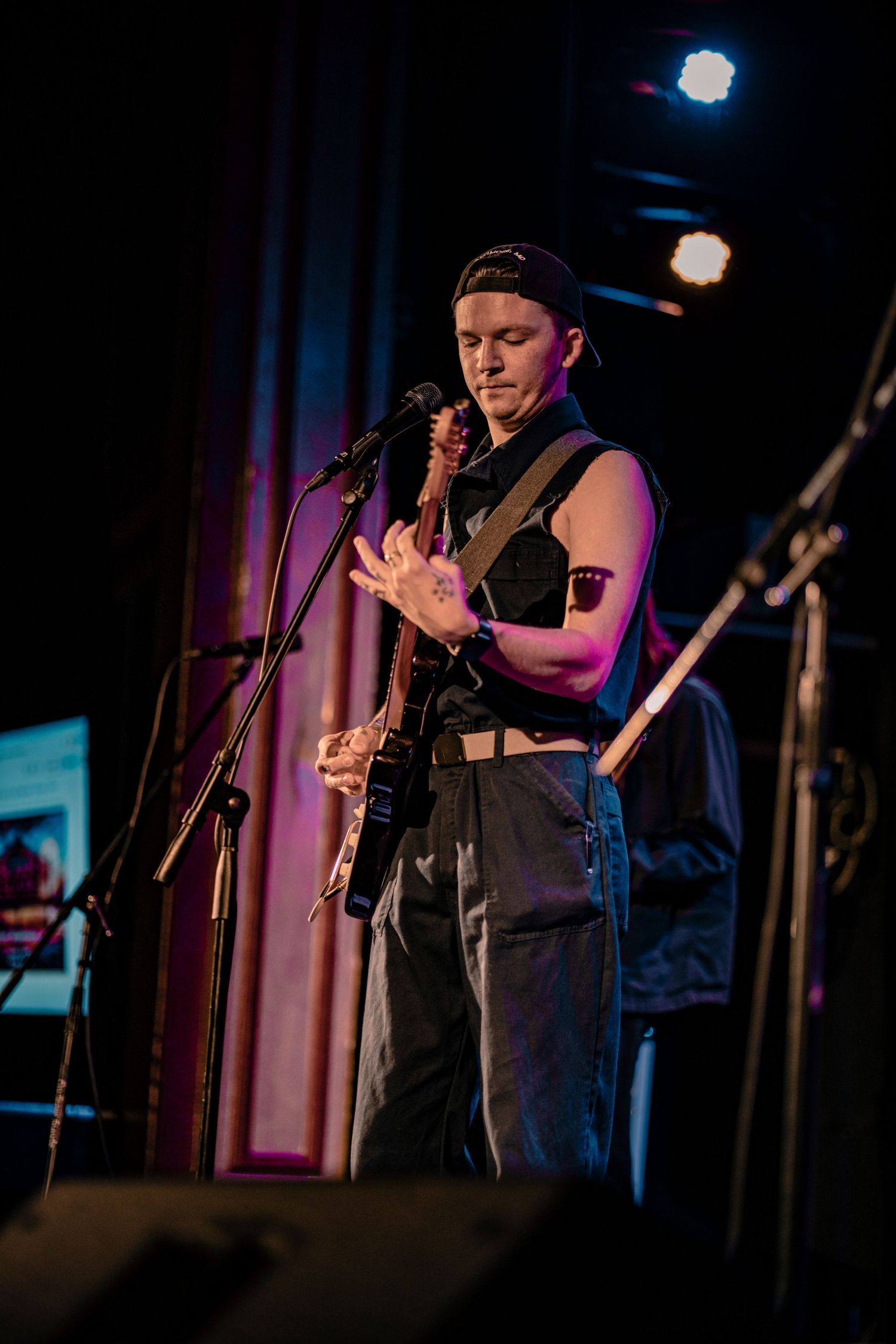 Musician in dark attire performs with guitar on stage under moody purple lighting.
