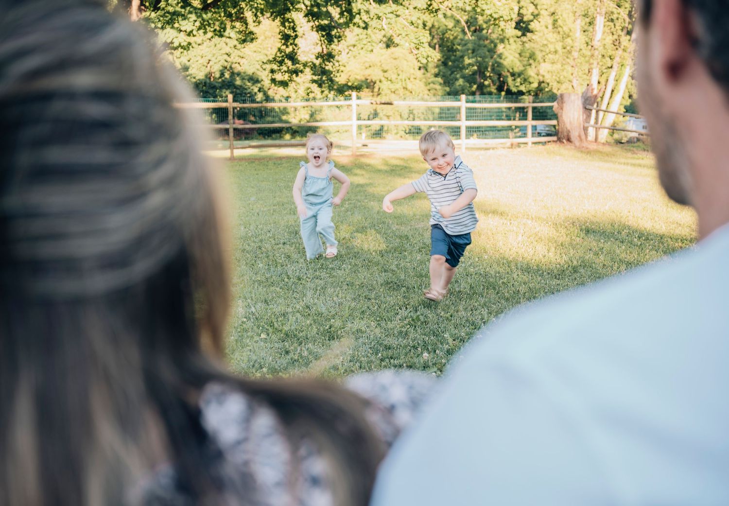 Two toddlers running toward parents on a sunny green lawn with trees and a wooden fence in the background.
