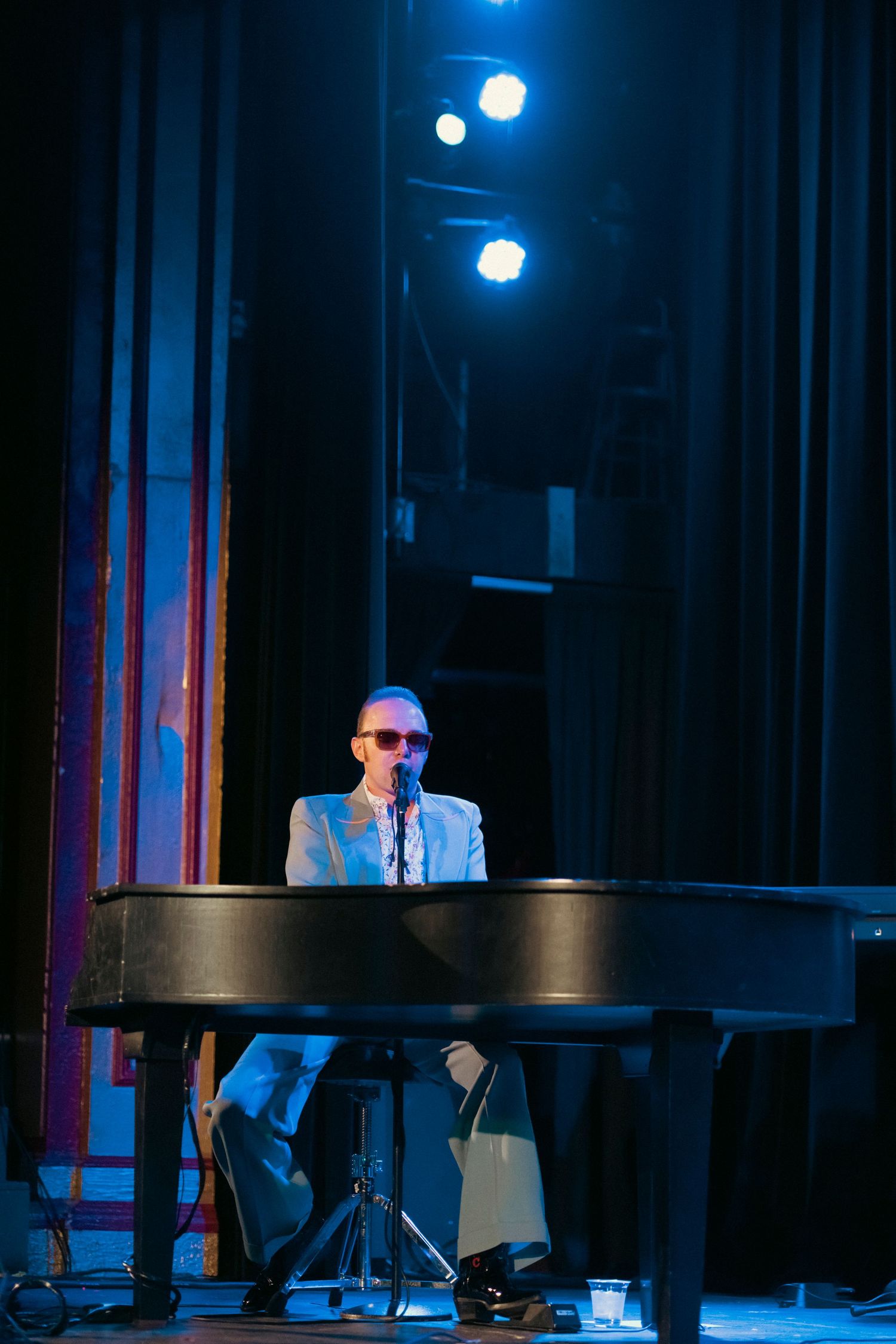 A performer in a light colored suit seated at a piano on a dimly lit stage.