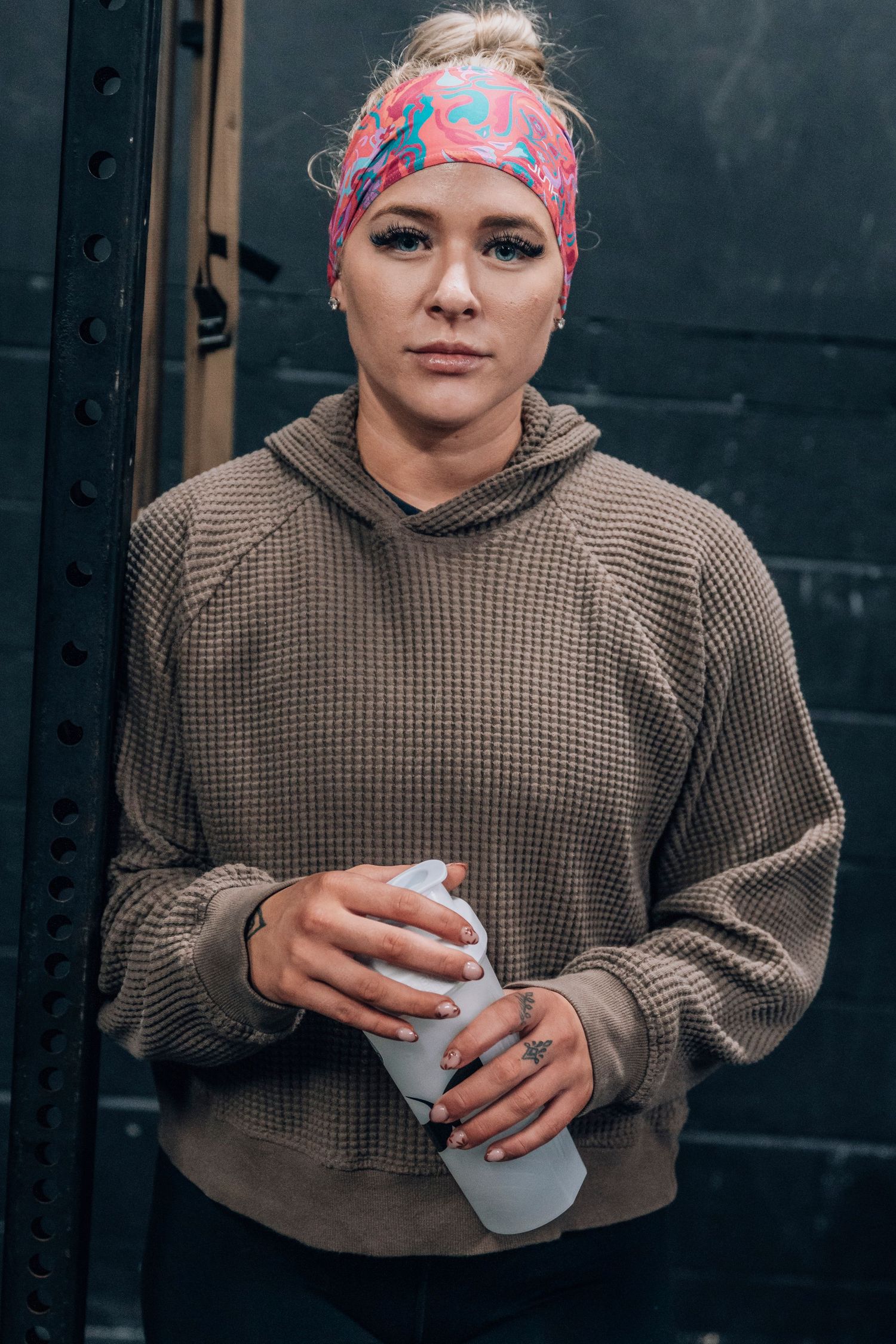 Athlete in brown hoodie and patterned headband holds water bottle against dark wall.