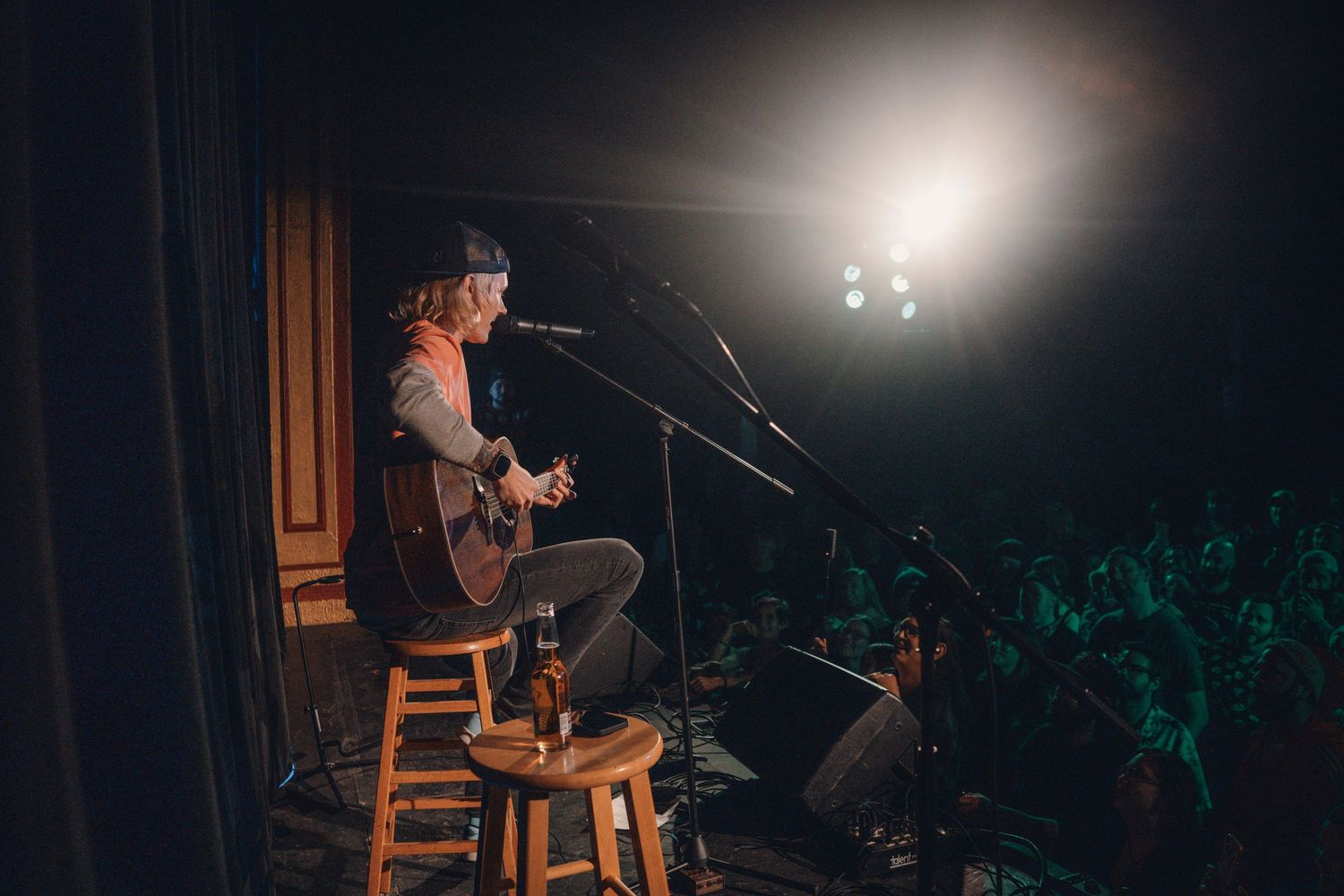 Performer seated on wooden stool under spotlight during acoustic set.