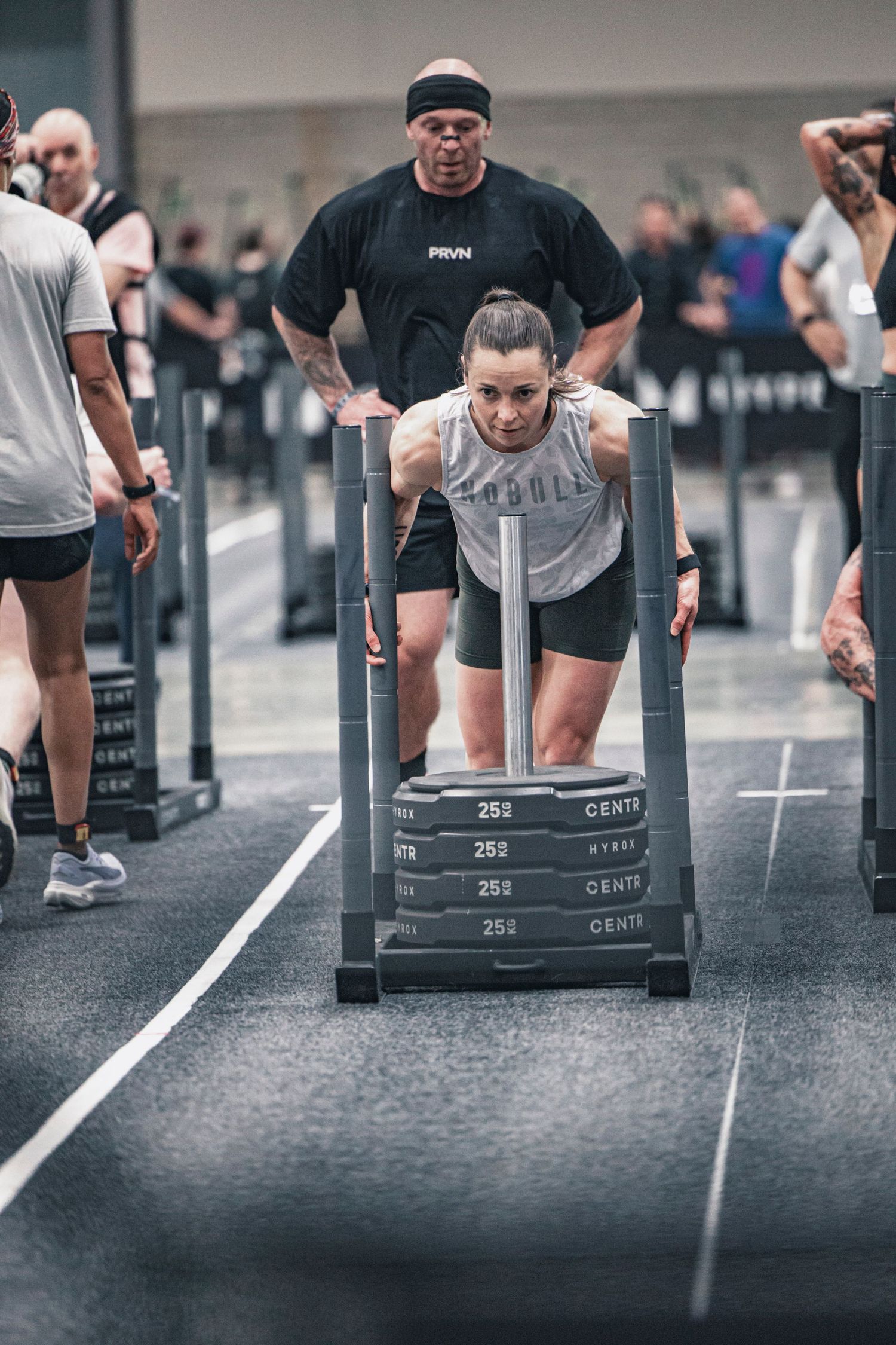Multiple athletes perform sled push exercises on a gym floor with numbered markers.