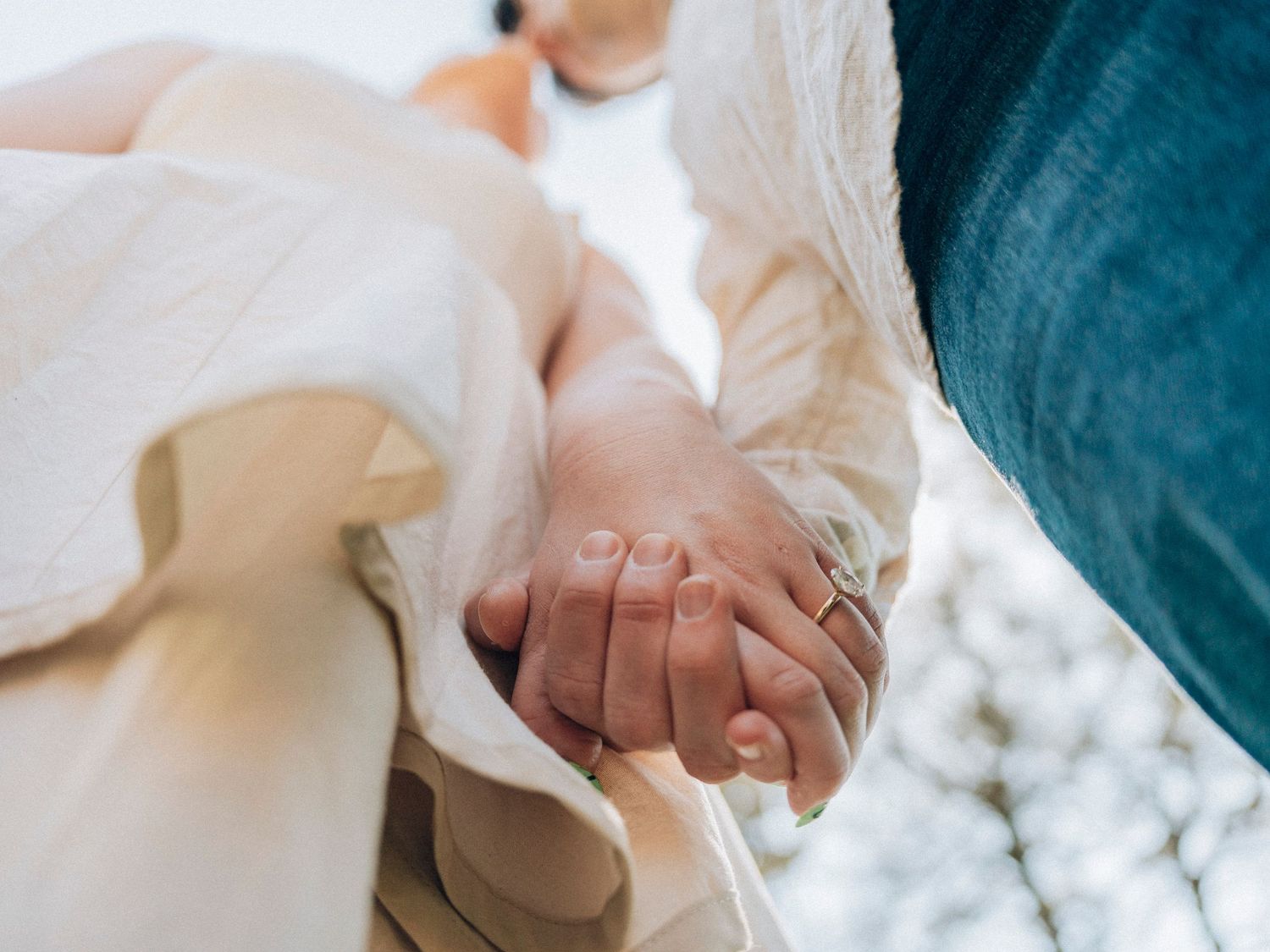 Close-up of a couple holding hands tenderly, with the woman wearing a flowing white dress outdoors.