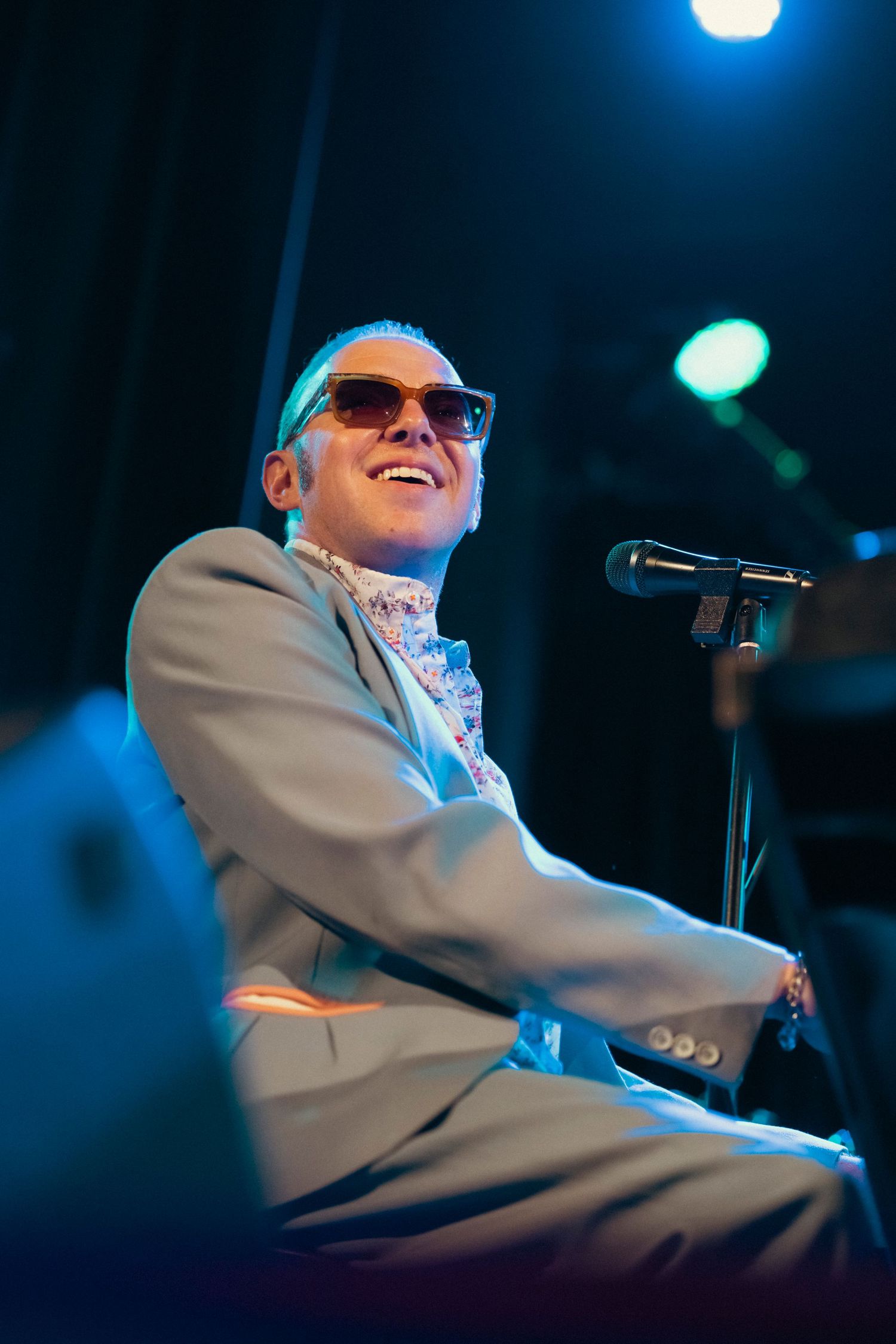 A performer in a light suit jacket playing piano under blue stage lighting.