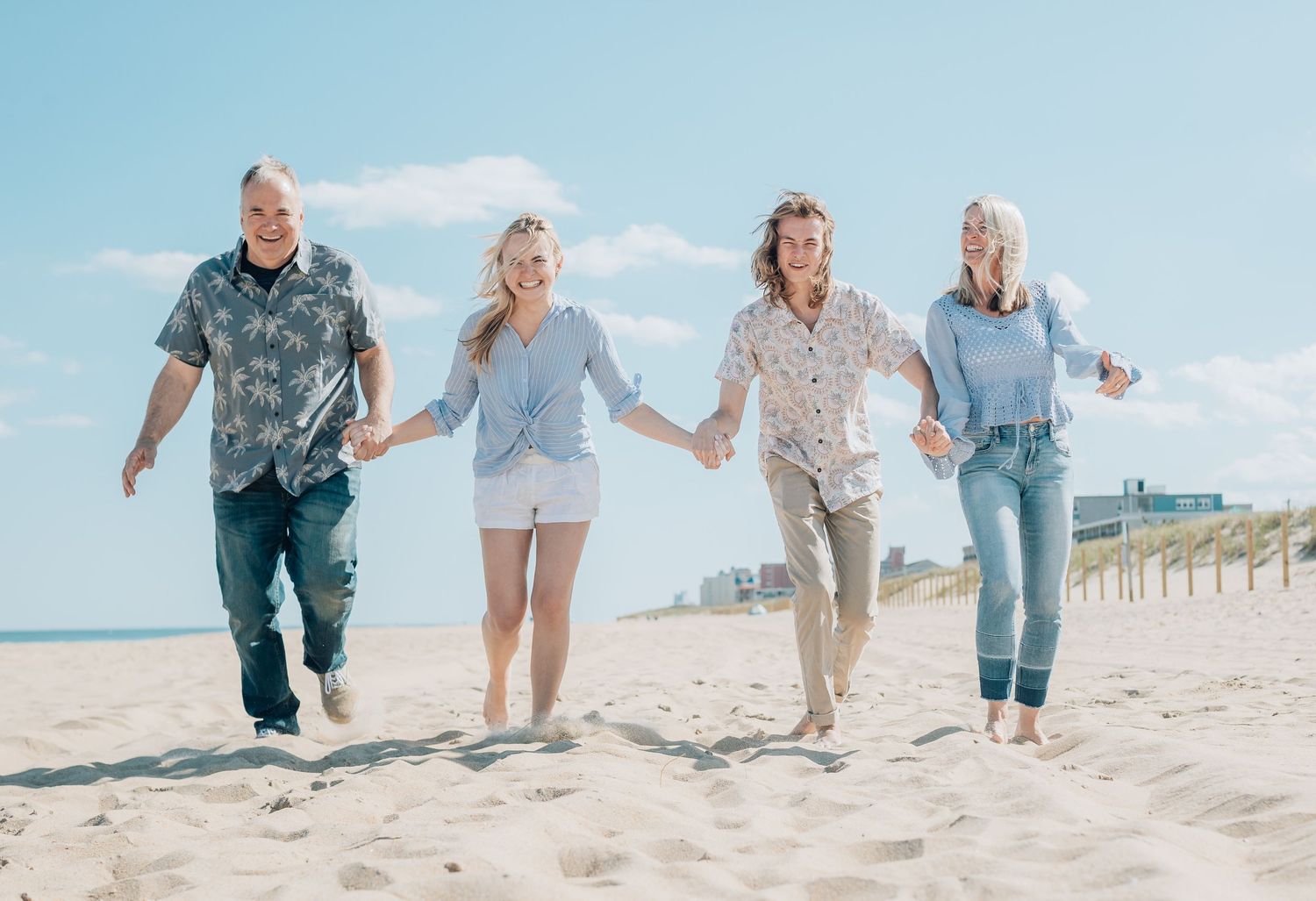 Four people holding hands while running and playing together on a sunny beach with white sand and blue sky.
