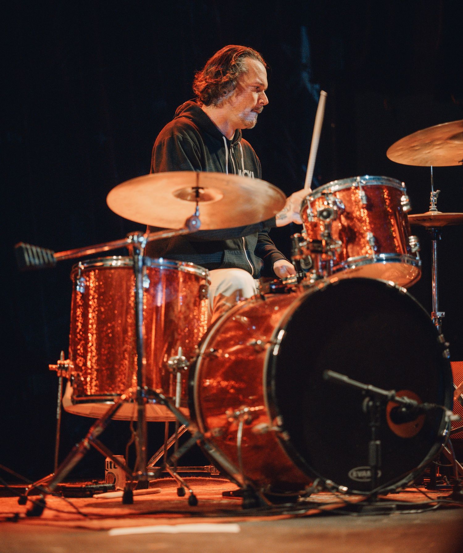 Orange-tinted drum kit setup on stage with multiple toms and cymbals under dramatic lighting.