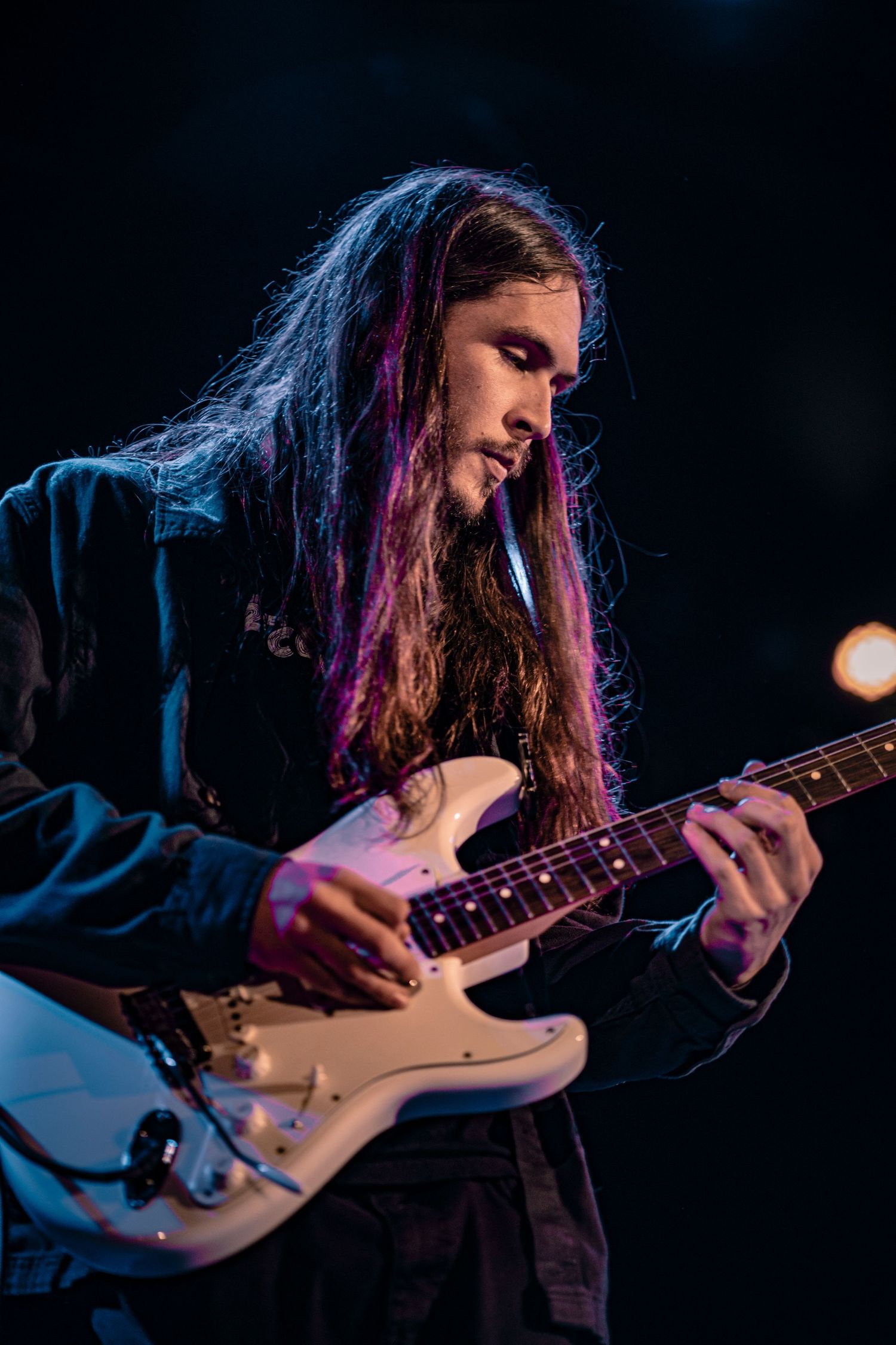 Guitarist with long hair plays electric guitar on dimly lit stage with blue lighting effects.