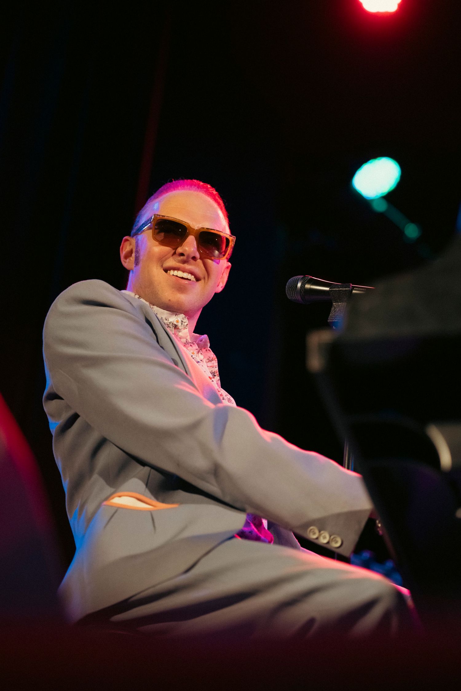 A performer in a grey suit and sunglasses sits at a piano on stage under colorful lighting.