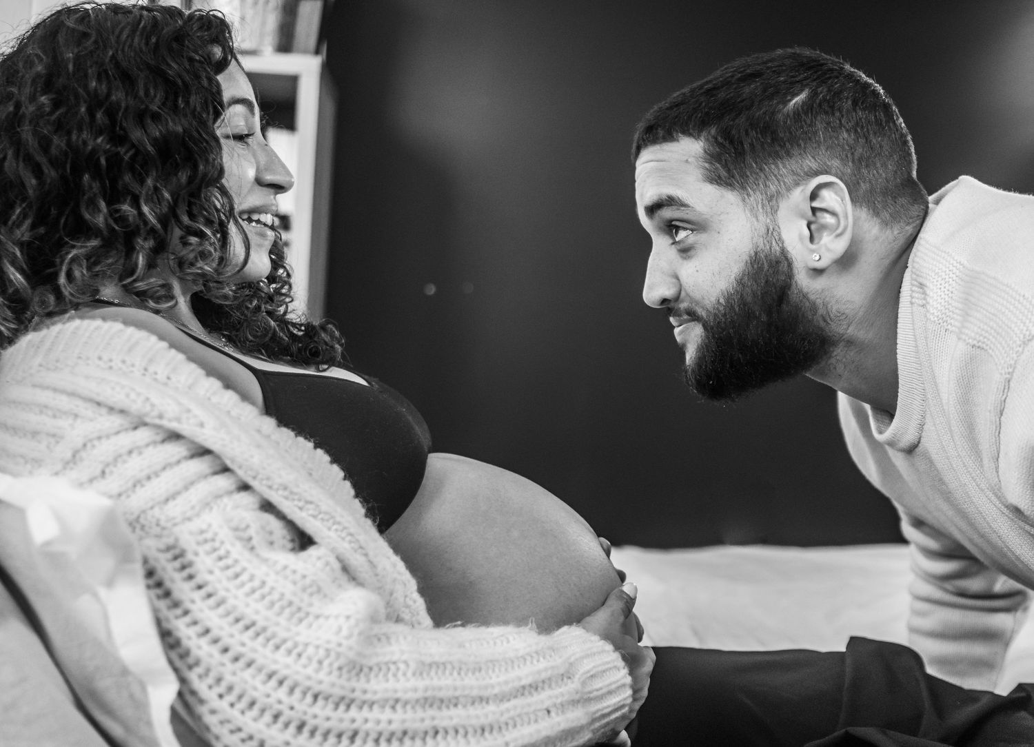 Black and white maternity photo of a bearded man smiling at pregnant woman's belly during intimate moment.