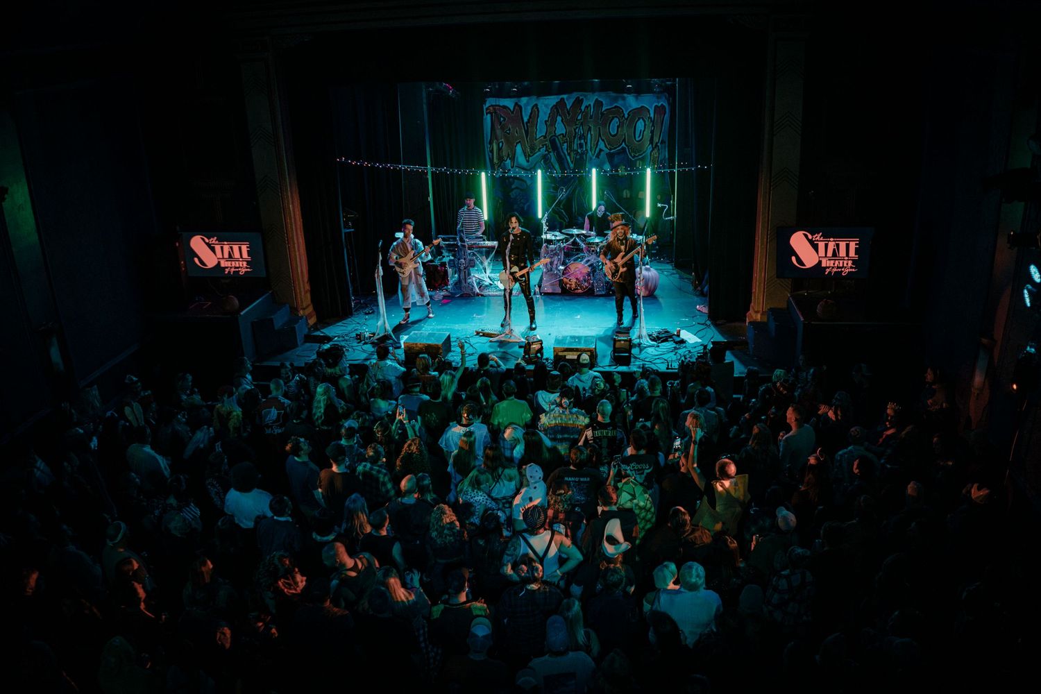Wide shot of concert venue with crowd and stage lighting in dark blue tones.