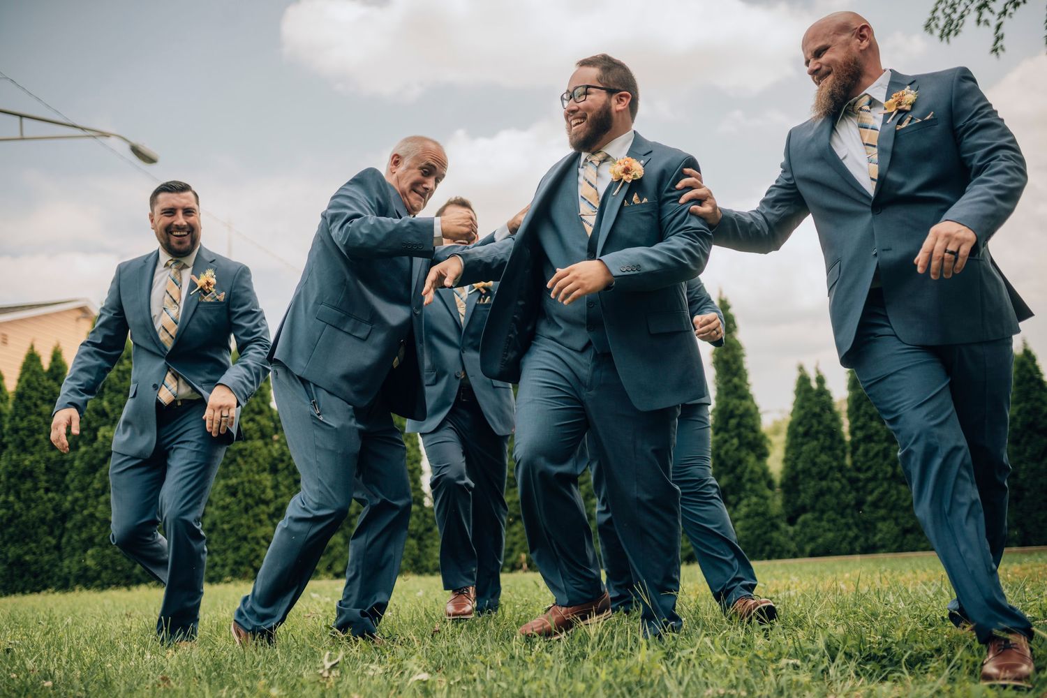 Groomsmen in blue suits strike playful poses on a lawn during wedding celebrations.