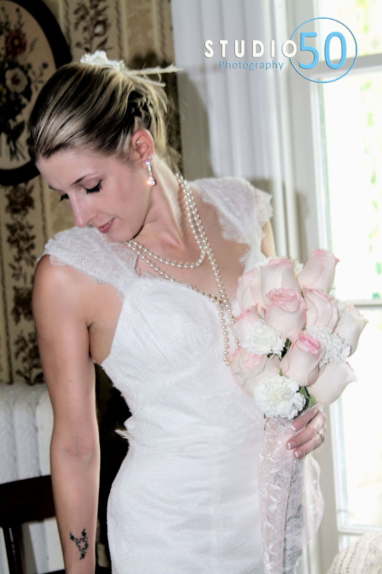 Elegant bride in white lace dress with pearl necklace holding pink and white bouquet.