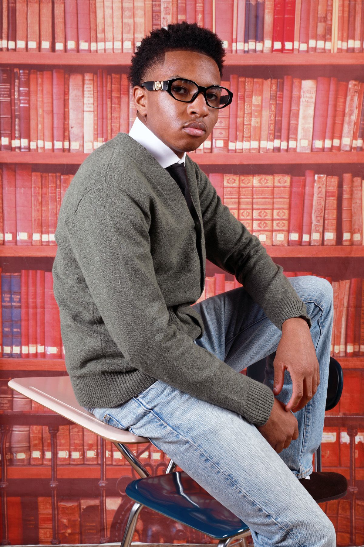 Academic portrait in library with classic sweater and glasses against red bookshelf background.