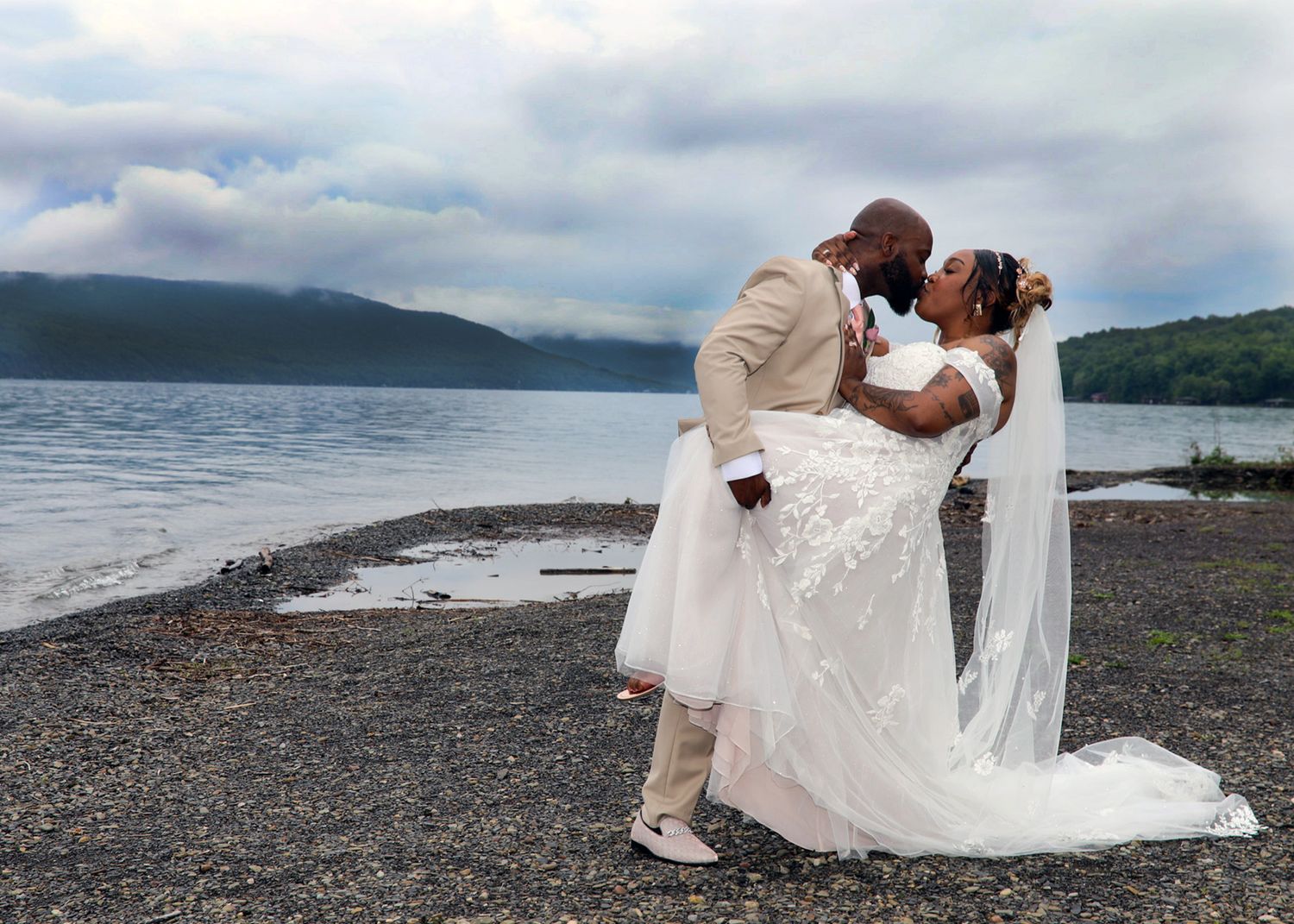 A couple shares a romantic kiss on a rocky beach shore with mountains and cloudy skies in the background.