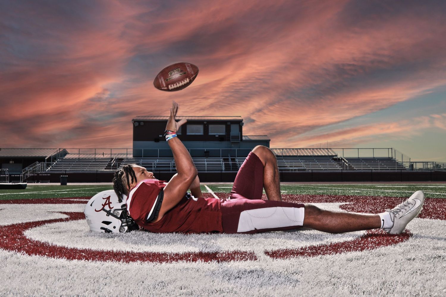 Football player lying on field holds ball up against dramatic sunset sky.