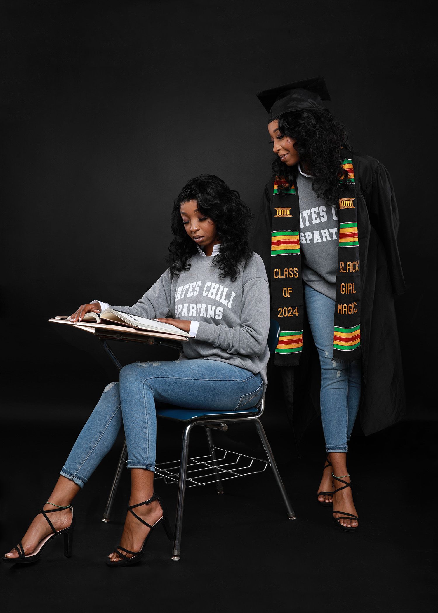 Two graduates in casual attire pose against dark backdrop with books and academic stoles.