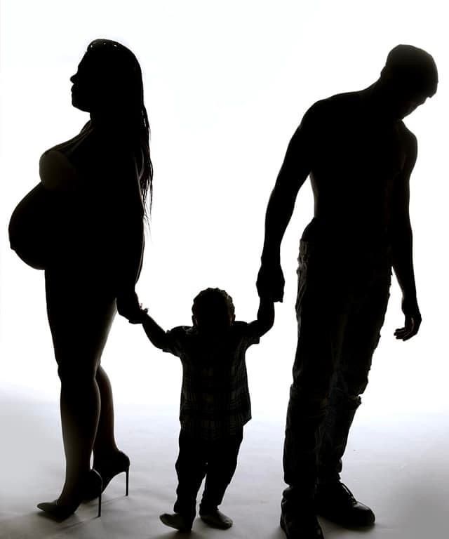 Artistic silhouette of pregnant family holding hands against white backdrop.