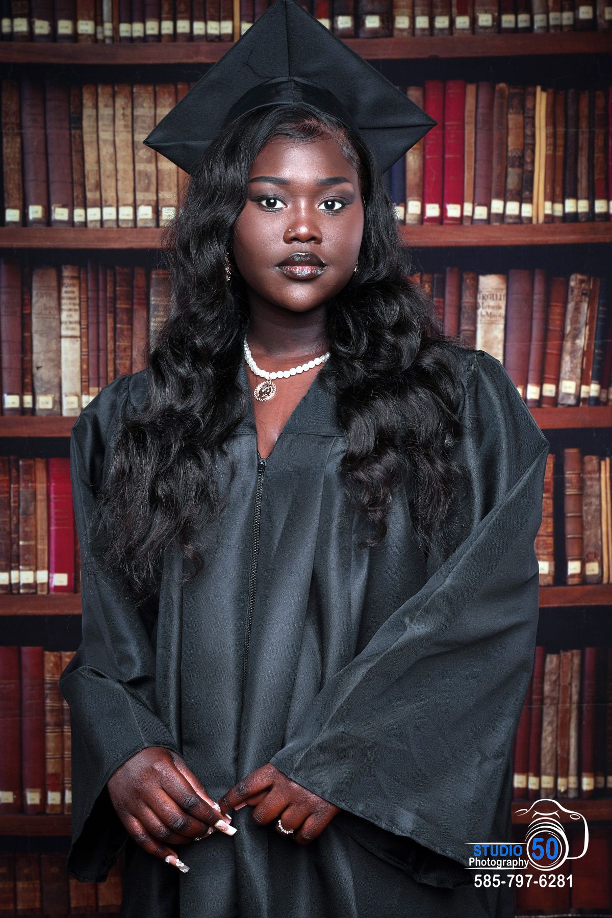 Graduate in black cap and gown poses in front of library bookshelves.
