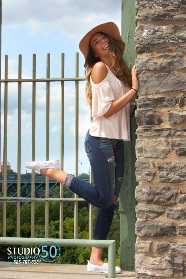 Senior portrait of woman in brown hat posing against stone wall and iron fence.