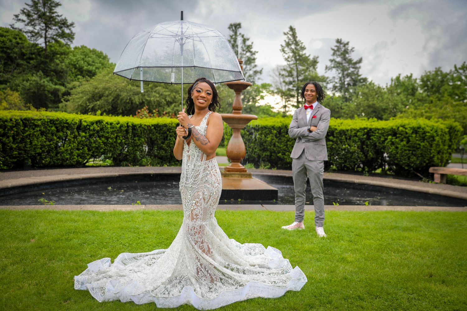 A joyful wedding moment on a lush green lawn with two people under an umbrella on a cloudy day.