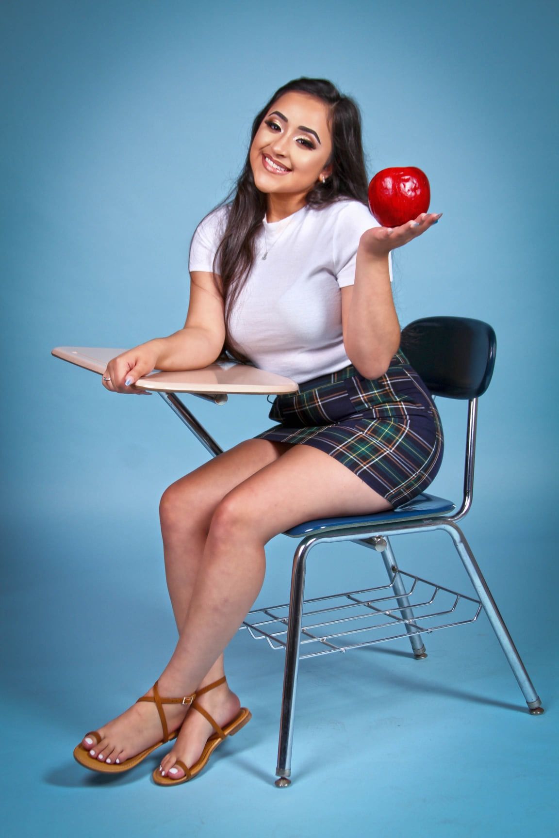 Student posing with an apple while sitting in a school chair against a blue backdrop for senior photos.
